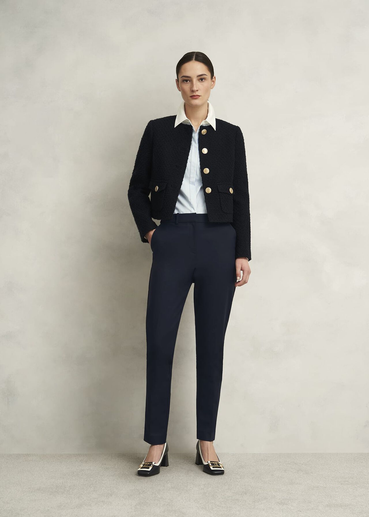 A woman wearing navy blue tapered trousers with a white shirt and black jacket, standing against a plain background.