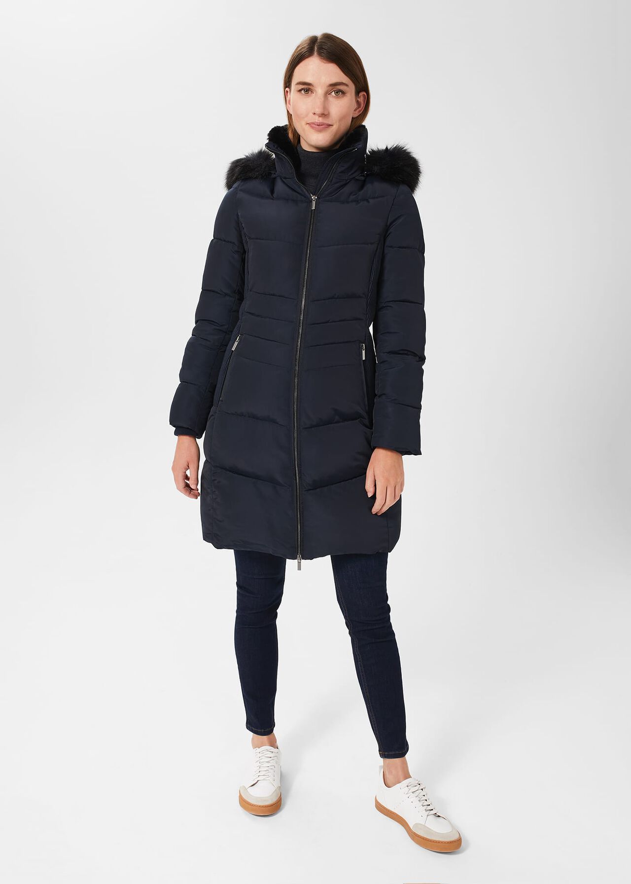 A woman wearing a navy blue hooded puffer coat with faux fur trim on the hood, front zip closure, and quilted pattern, paired with dark jeans and white sneakers.