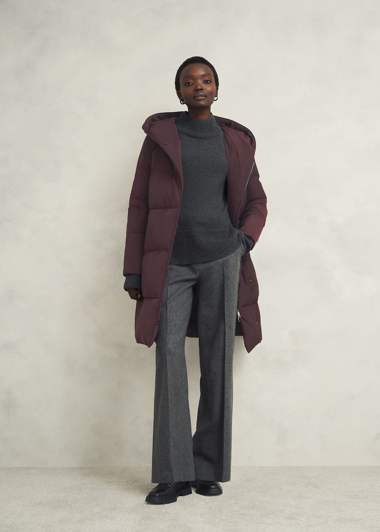 A woman wearing a long maroon hooded puffer jacket with a front zip, paired with grey trousers and a dark grey sweater.