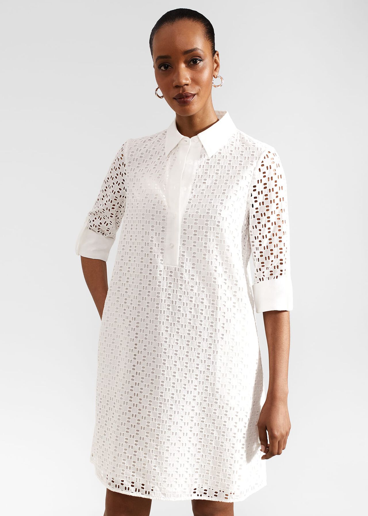 A woman wearing a white eyelet cotton dress with a collar, long rolled-up sleeves, and a button-down front.