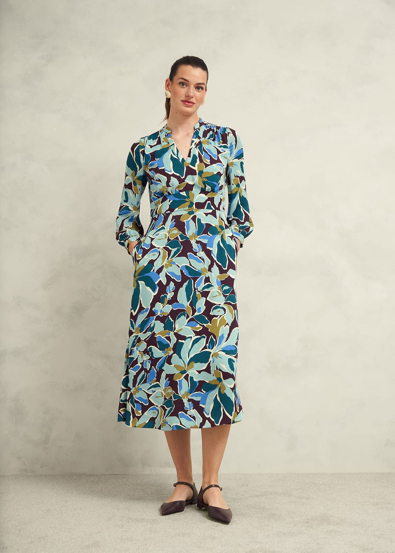 A woman wearing a colorful floral midi dress with a mandarin collar and long sleeves, standing against a plain background.