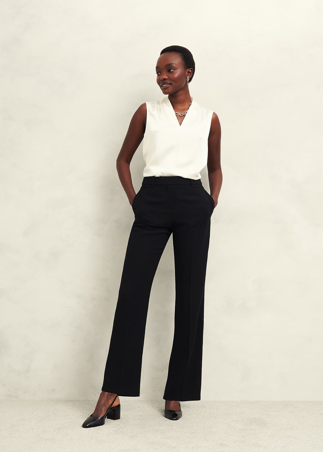 A woman wearing black straight trousers with a white sleeveless top, standing against a plain background.