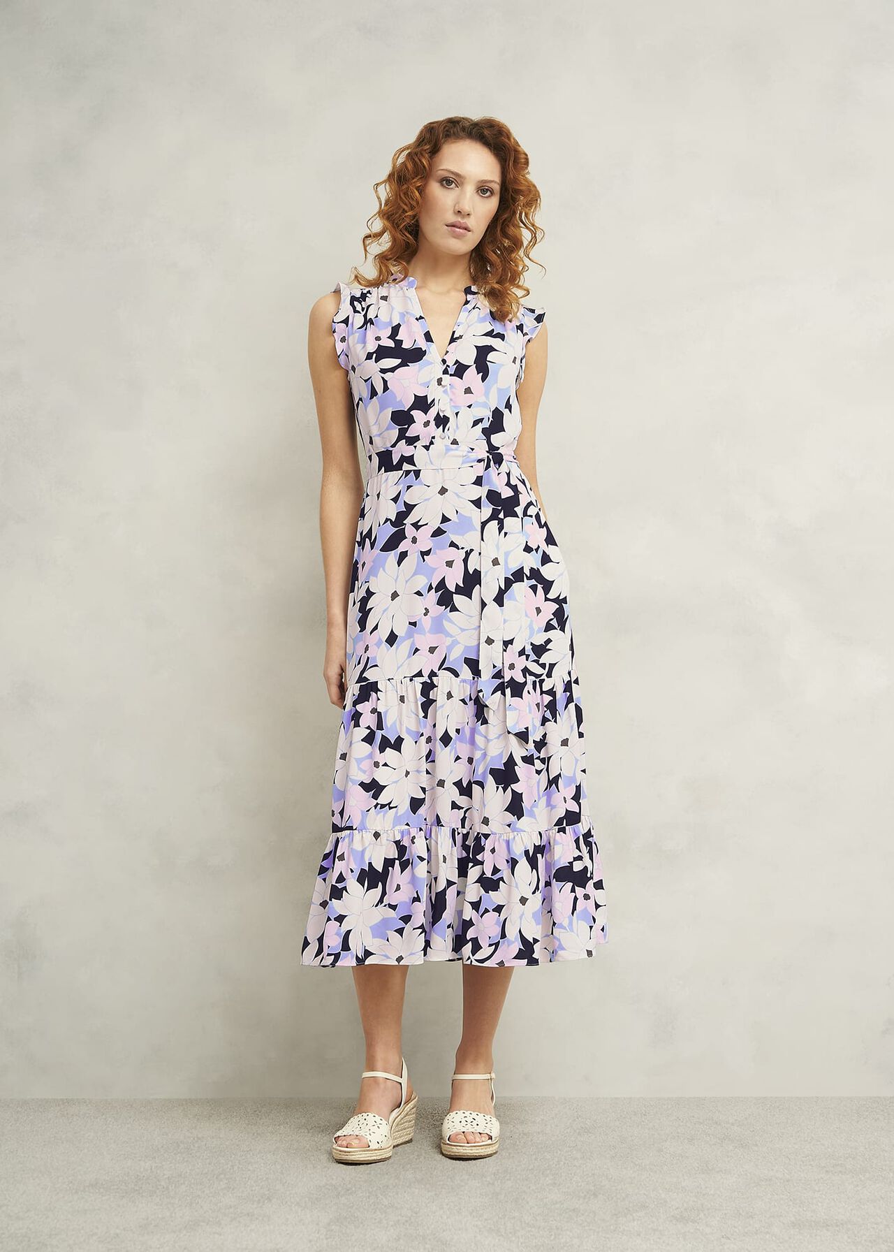 A woman wearing a sleeveless, floral midi dress with a mandarin collar, side zip, and tiered skirt, in shades of white, black, and pastel purple.