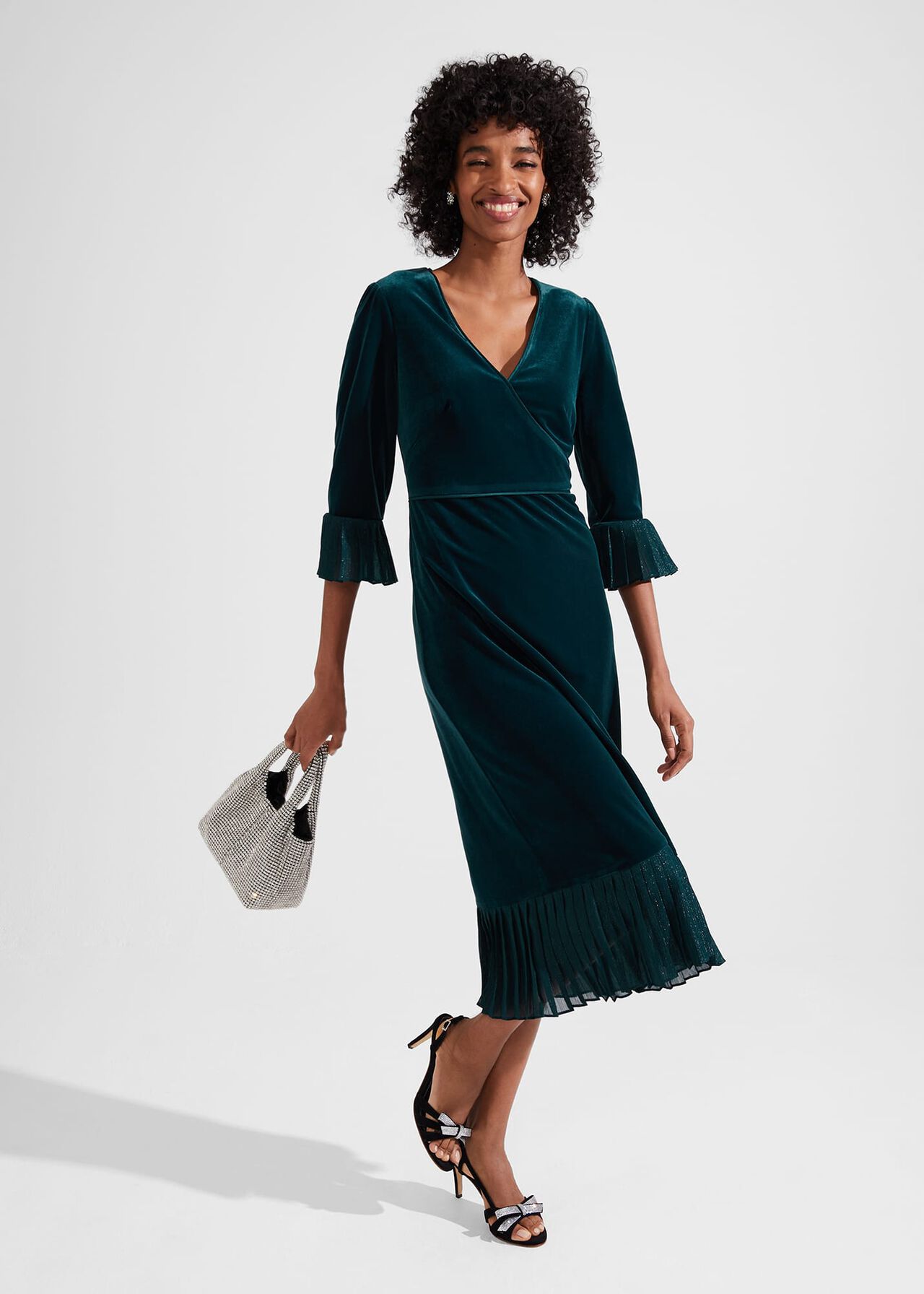 A woman wearing a teal velvet midi dress with a V-neckline, long pleated cuffs on the sleeves, and a pleated hem, holding a small handbag and wearing black high heels.