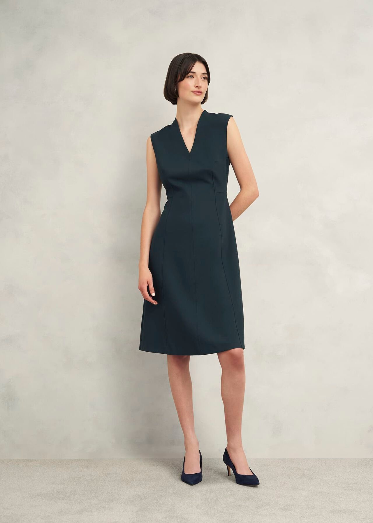 A woman wearing a navy blue sleeveless dress with a V-neckline, fitted waist, and knee-length hem, paired with navy high heels.