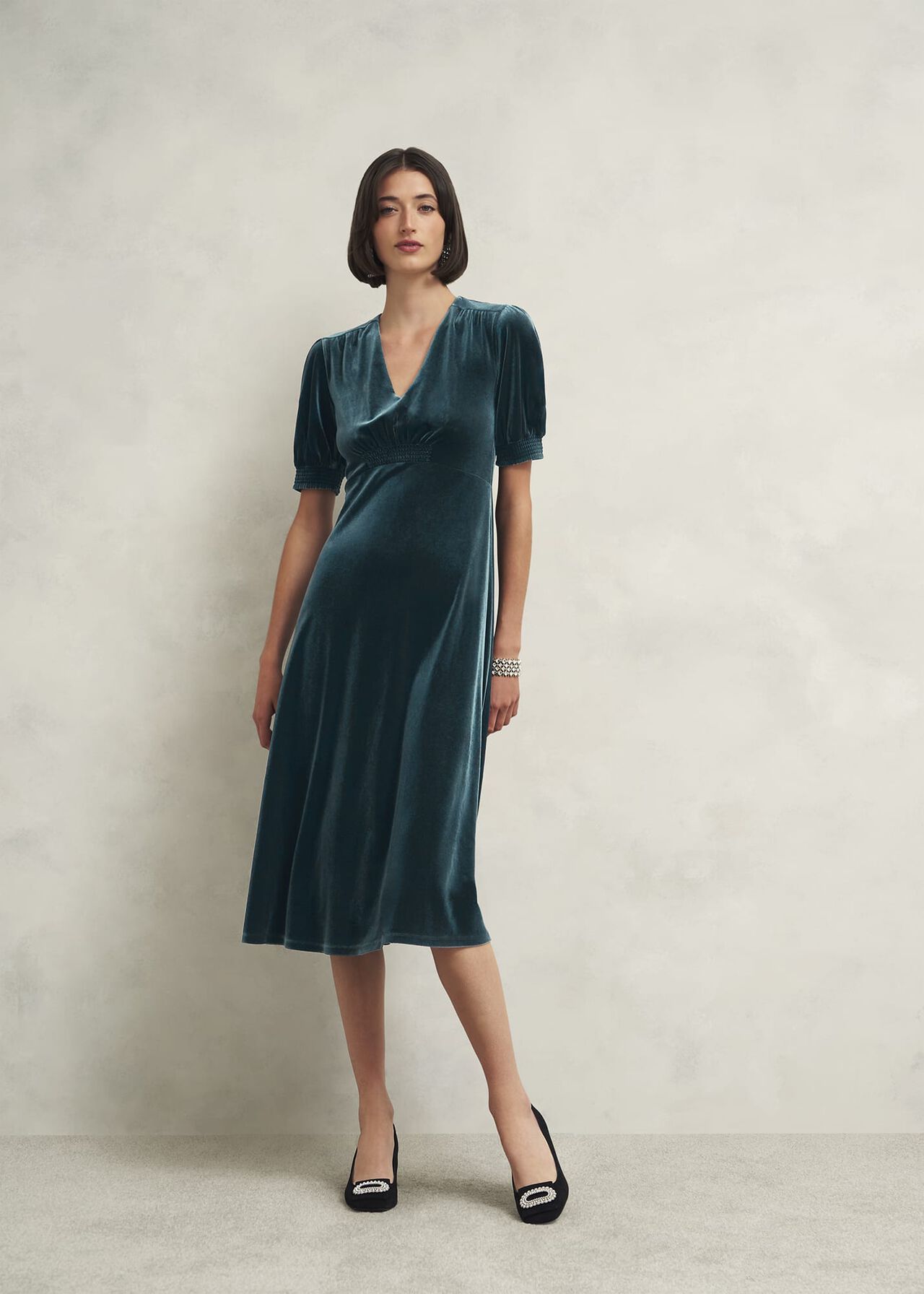 A woman wearing a deep teal velvet dress with a V-neckline, short puffed sleeves, fitted waist, and midi length, standing against a plain background.