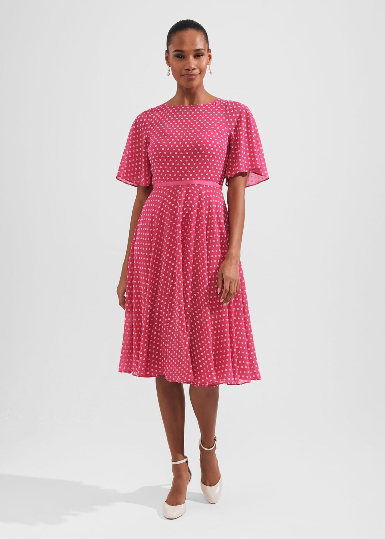 A woman wearing a pink spot fit and flare dress with short flutter sleeves, crew neckline, and a gathered waist, standing against a plain background.