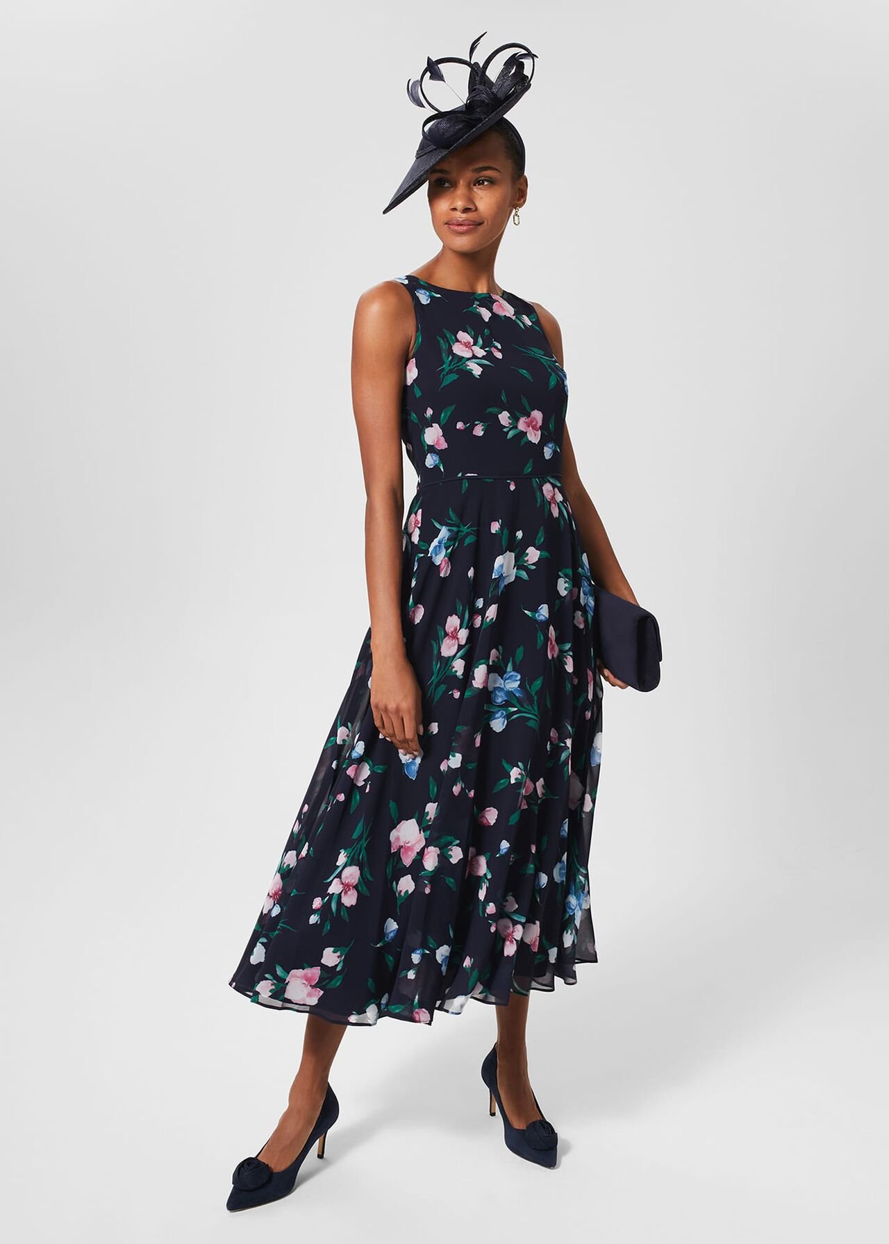 A woman wearing a navy floral dress with pink, white, and blue flowers, sleeveless with a crew neckline, paired with navy heels and a large navy fascinator.