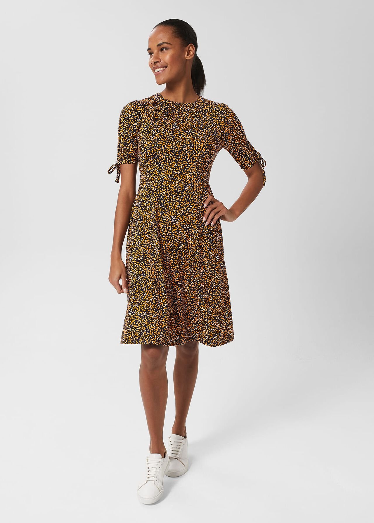 A woman wearing a knee-length dress with a multicolour abstract print, crew neckline, short sleeves with ties, and a fitted silhouette.