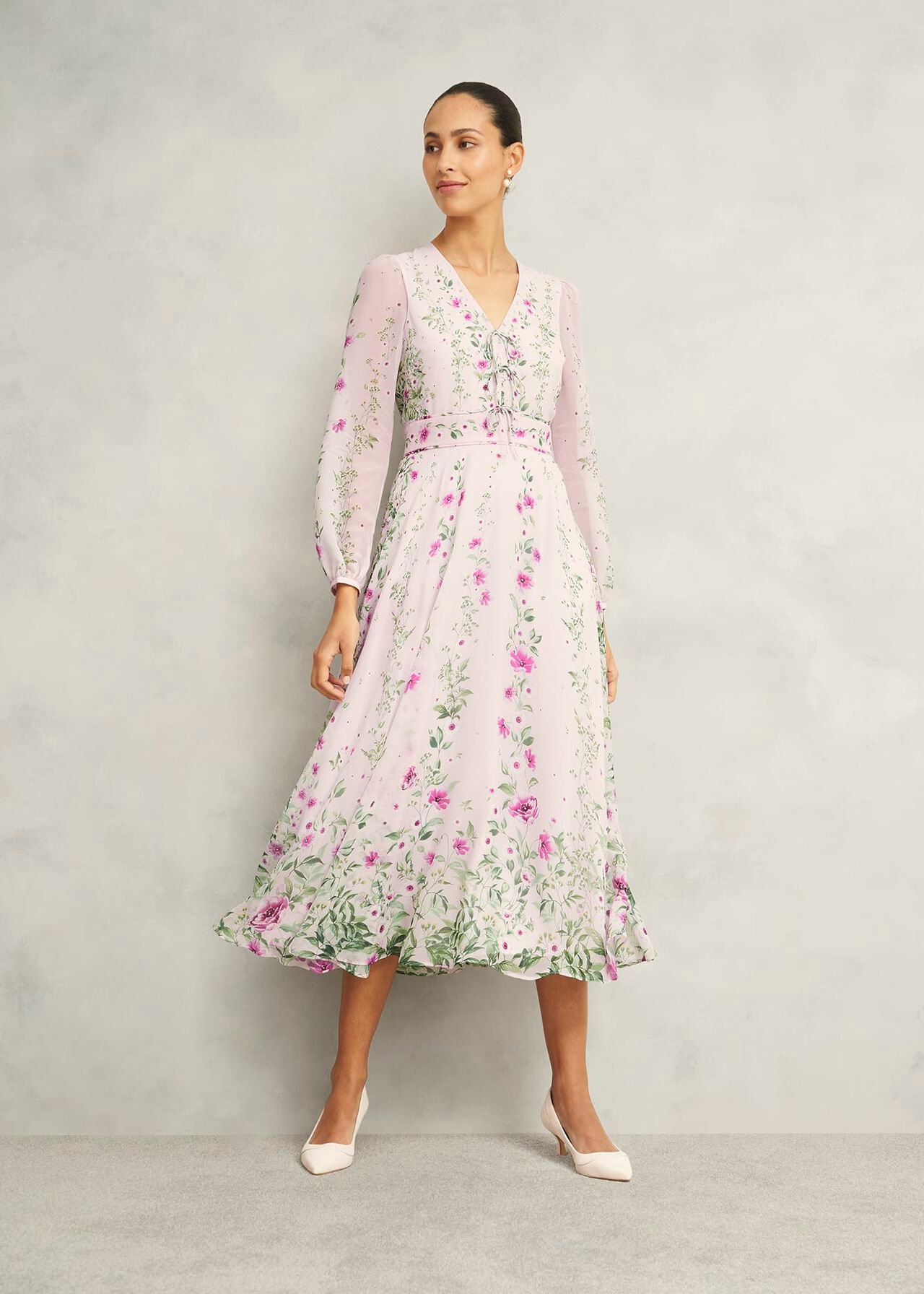A woman wearing a pink floral silk dress with a V-neckline, long sheer sleeves, and a bow at the waist, standing against a plain background.