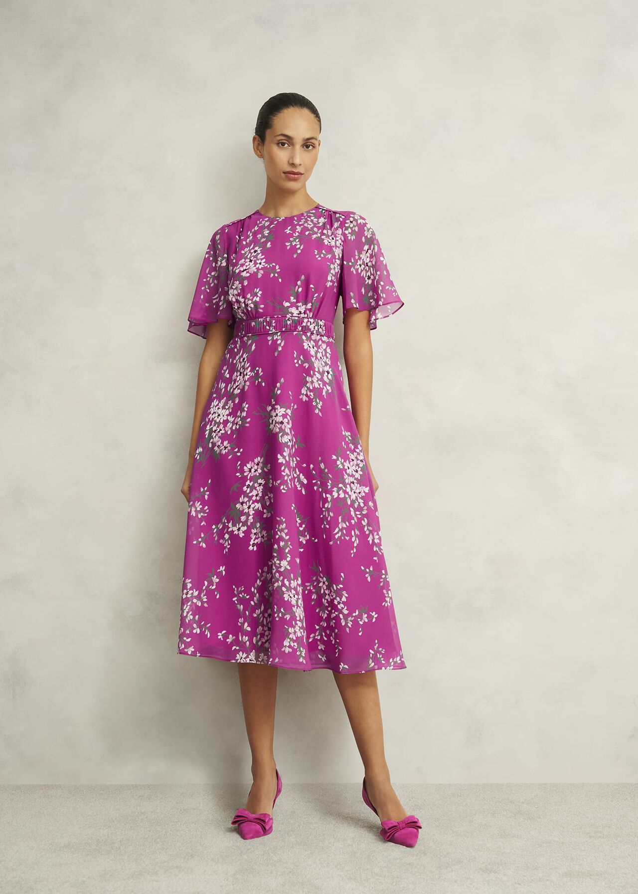 A woman wearing a pink floral silk dress with short flutter sleeves, a crew neckline, and a midi length, standing against a plain background.