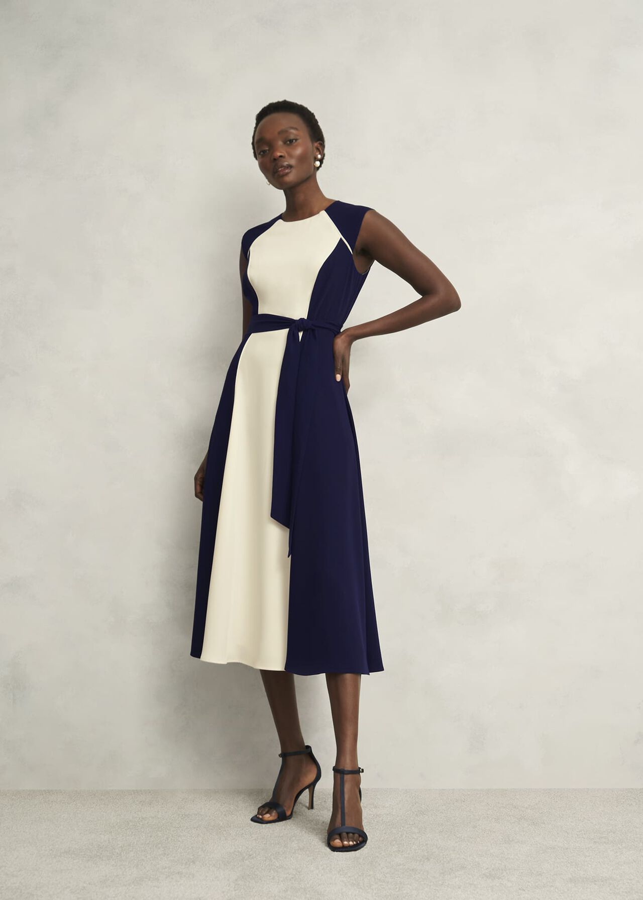 A woman wearing a navy and cream colourblock dress with sleeveless design, crew neckline, and a waist tie, paired with black high-heeled sandals.
