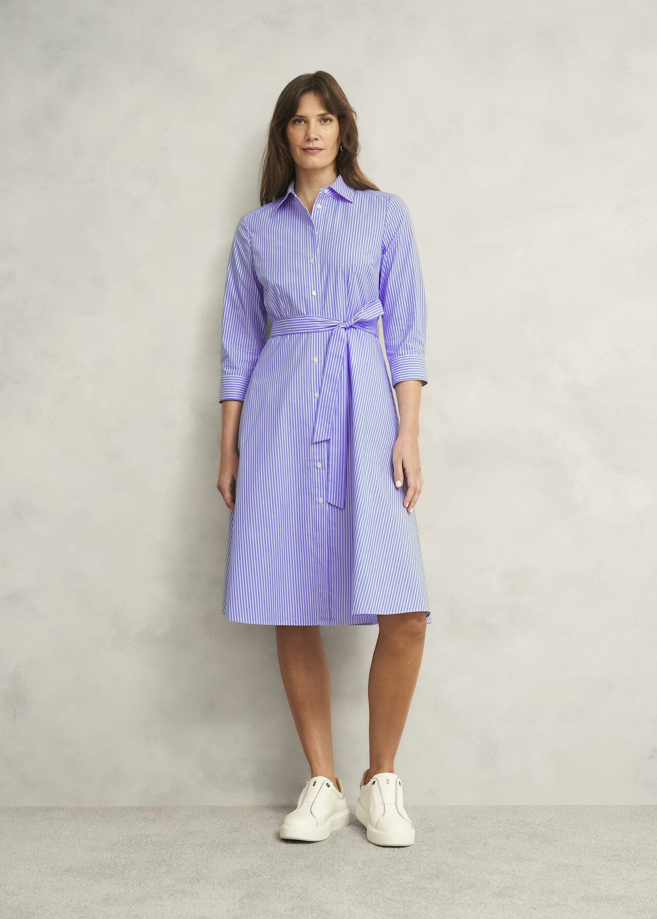 A woman wearing a blue and white striped shirt dress with a collar, front buttons, three-quarter sleeves, and a fabric belt, paired with white sneakers.
