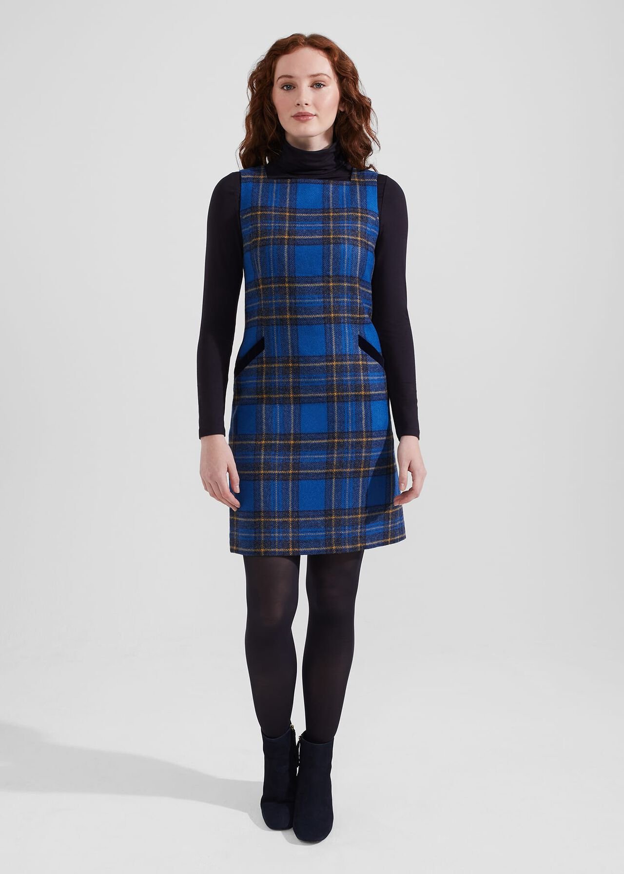 A woman wearing a blue tartan wool dress with a square neckline, sleeveless design, and a fitted silhouette, paired with black tights and ankle boots.