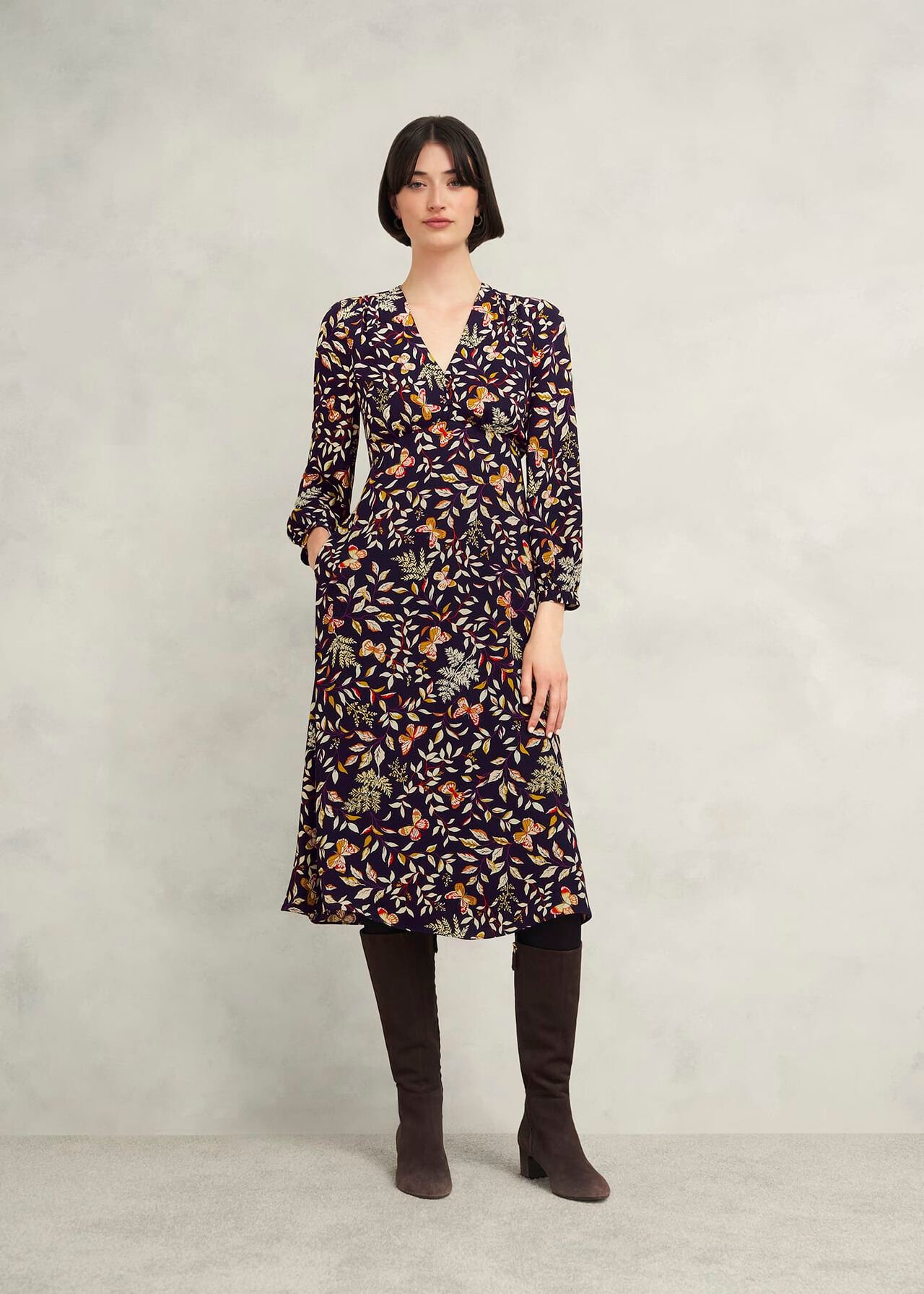 A woman wearing a navy blue floral dress with long sleeves, V-neckline, and a back zip fastening, paired with knee-high brown boots.
