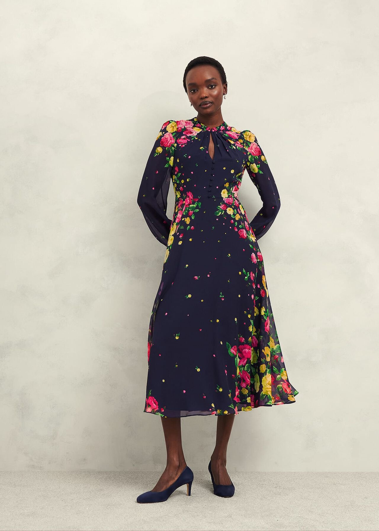 A woman wearing a navy blue floral silk dress with long sheer sleeves, a Mandarin collar, and a zip fastening, featuring a vibrant pink, yellow, and green floral pattern.