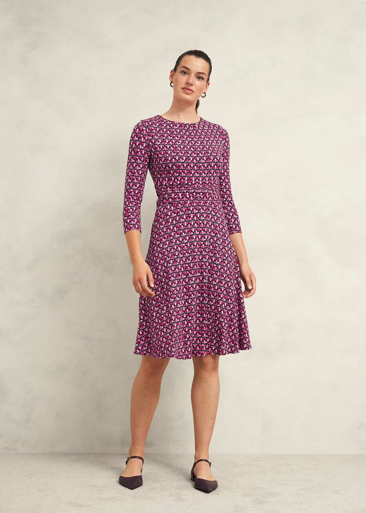 A woman wearing a pink, purple, and white patterned knee-length dress with a crew neckline and three-quarter sleeves, standing against a plain background.