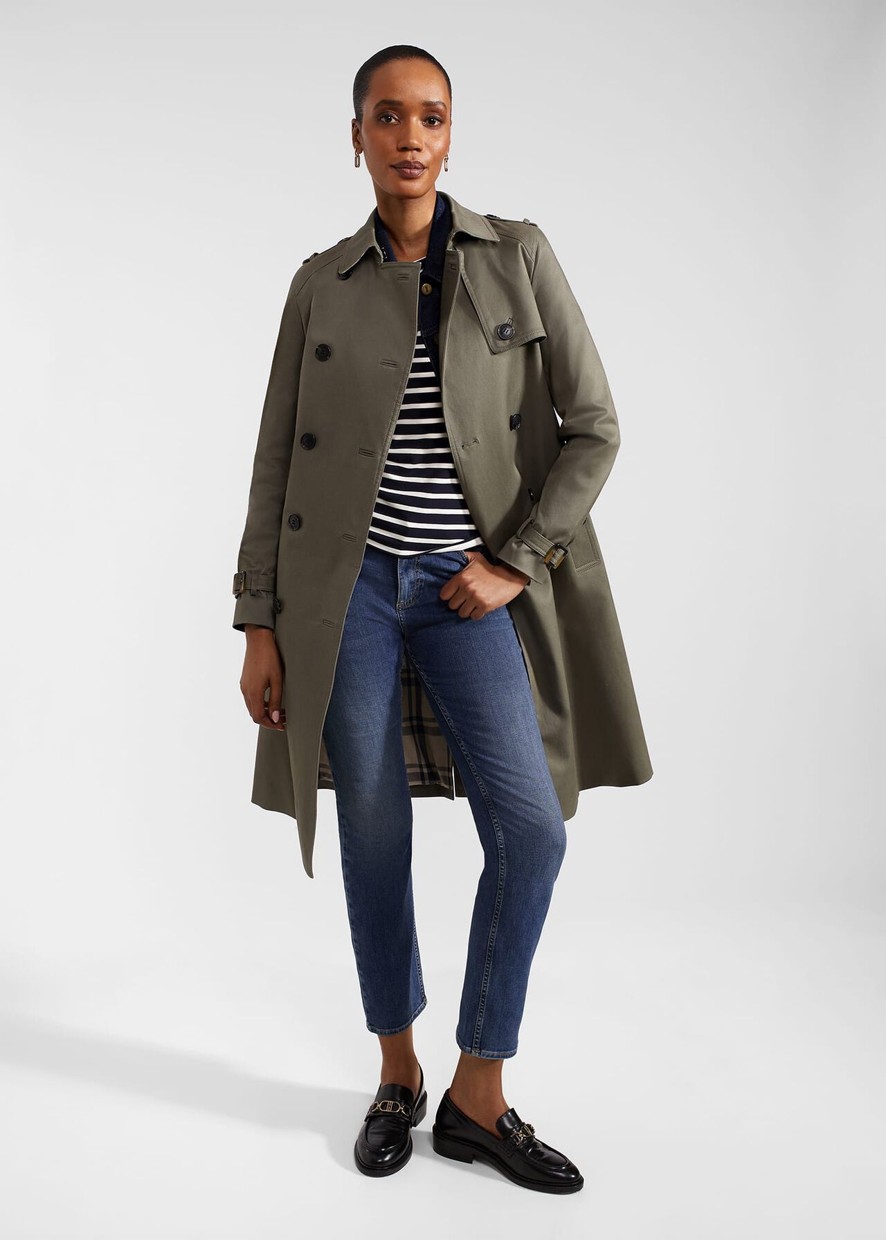 A woman wearing a beige trench coat with a collar, double-breasted front, and knee-length cut.