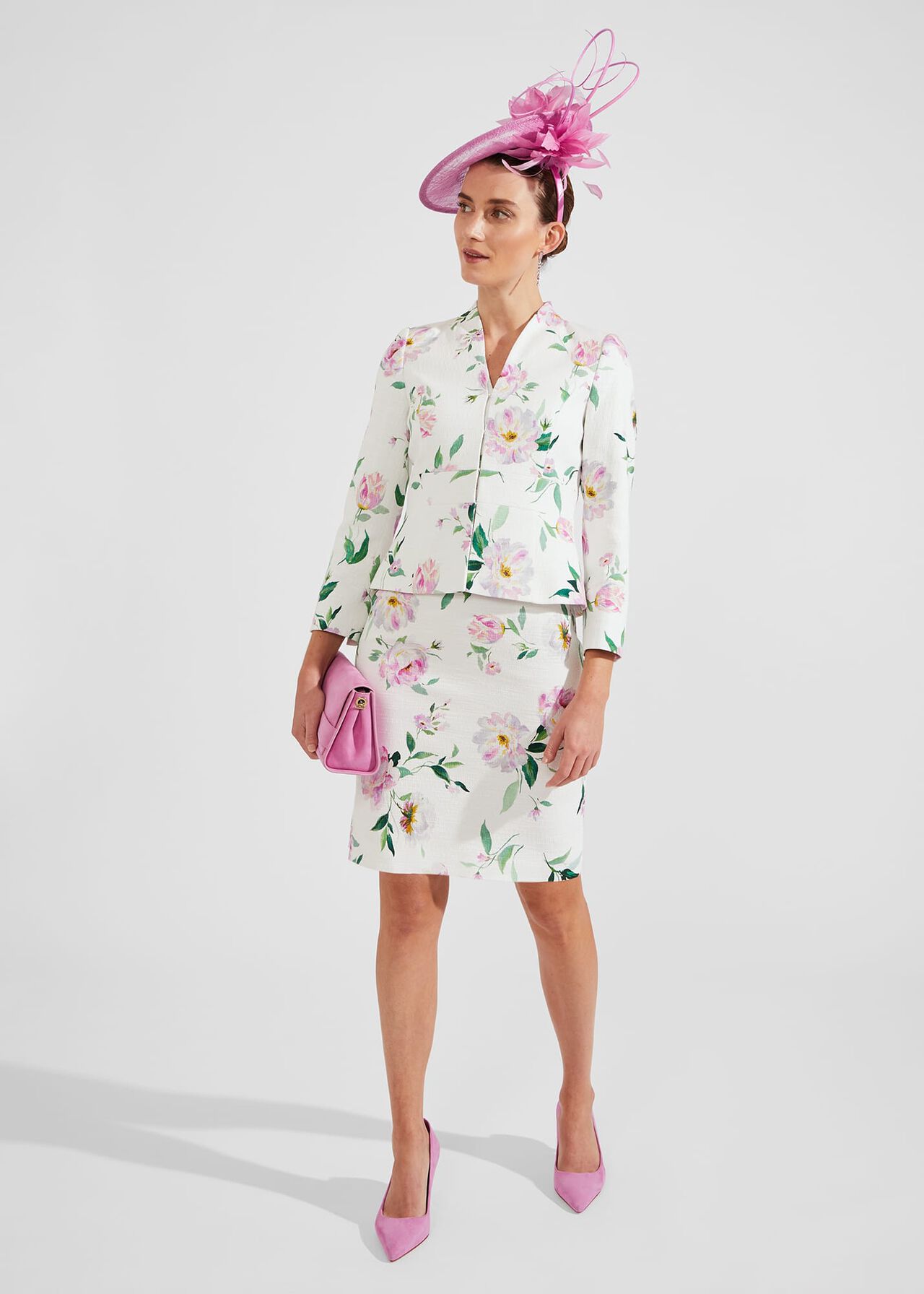 A woman wearing a cream floral tweed jacket with pink, green, and yellow flowers, collarless, front fastening, paired with a matching skirt, pink accessories, and pink heels.