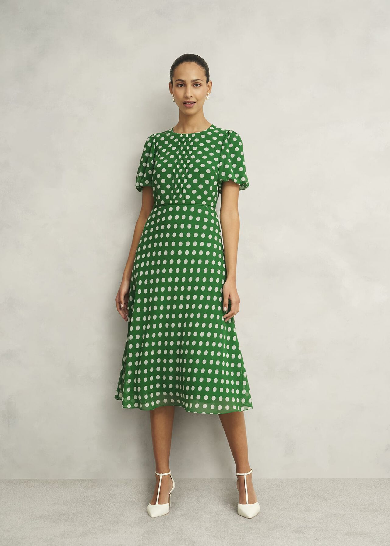 A woman wearing a green dress with white polka dots, short puffed sleeves, crew neckline, fitted waist, and midi length, standing against a plain background.