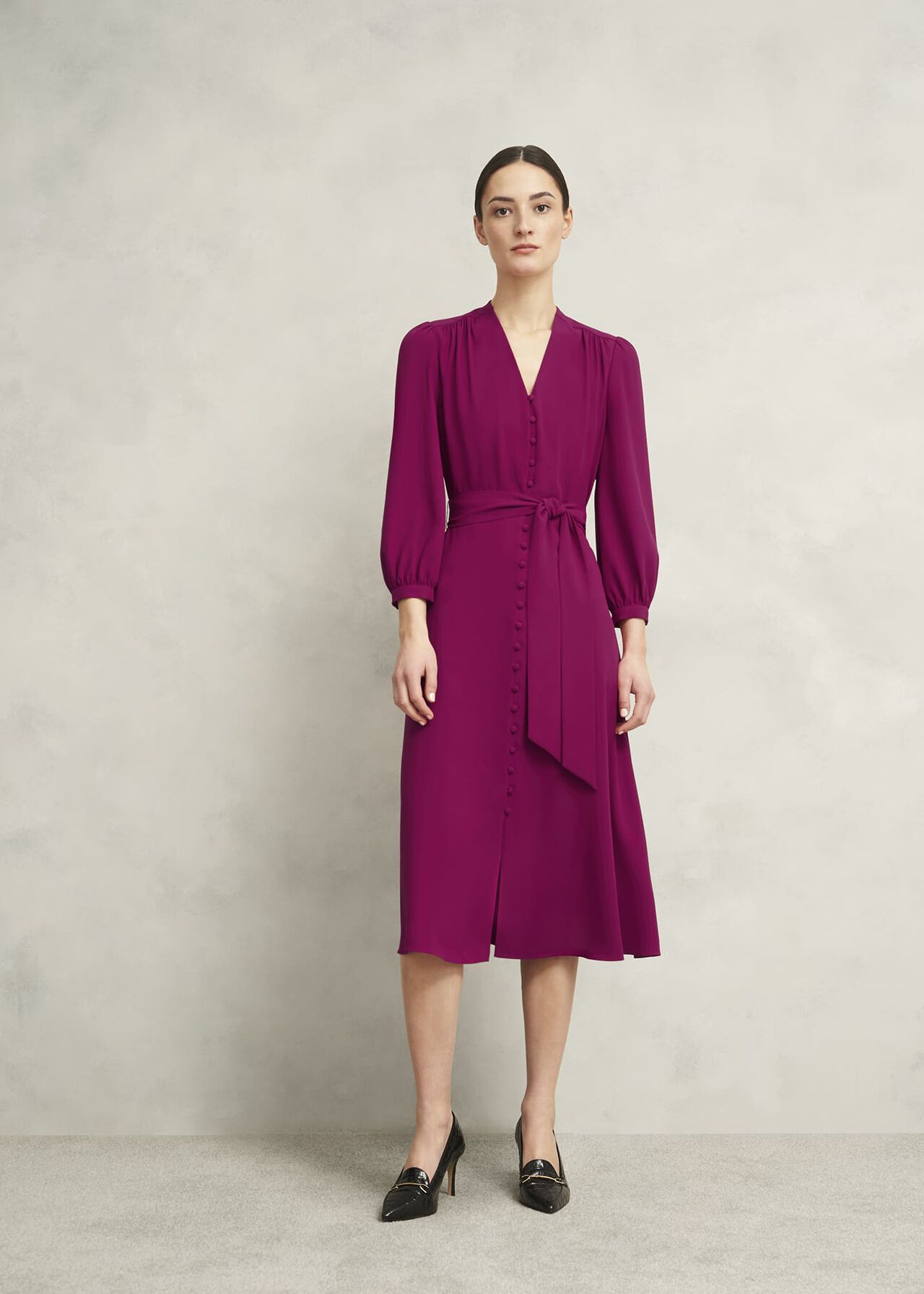 A woman wearing a magenta/pink midi dress with a V-neckline, long puffed sleeves, and a waist tie, standing against a plain background.
