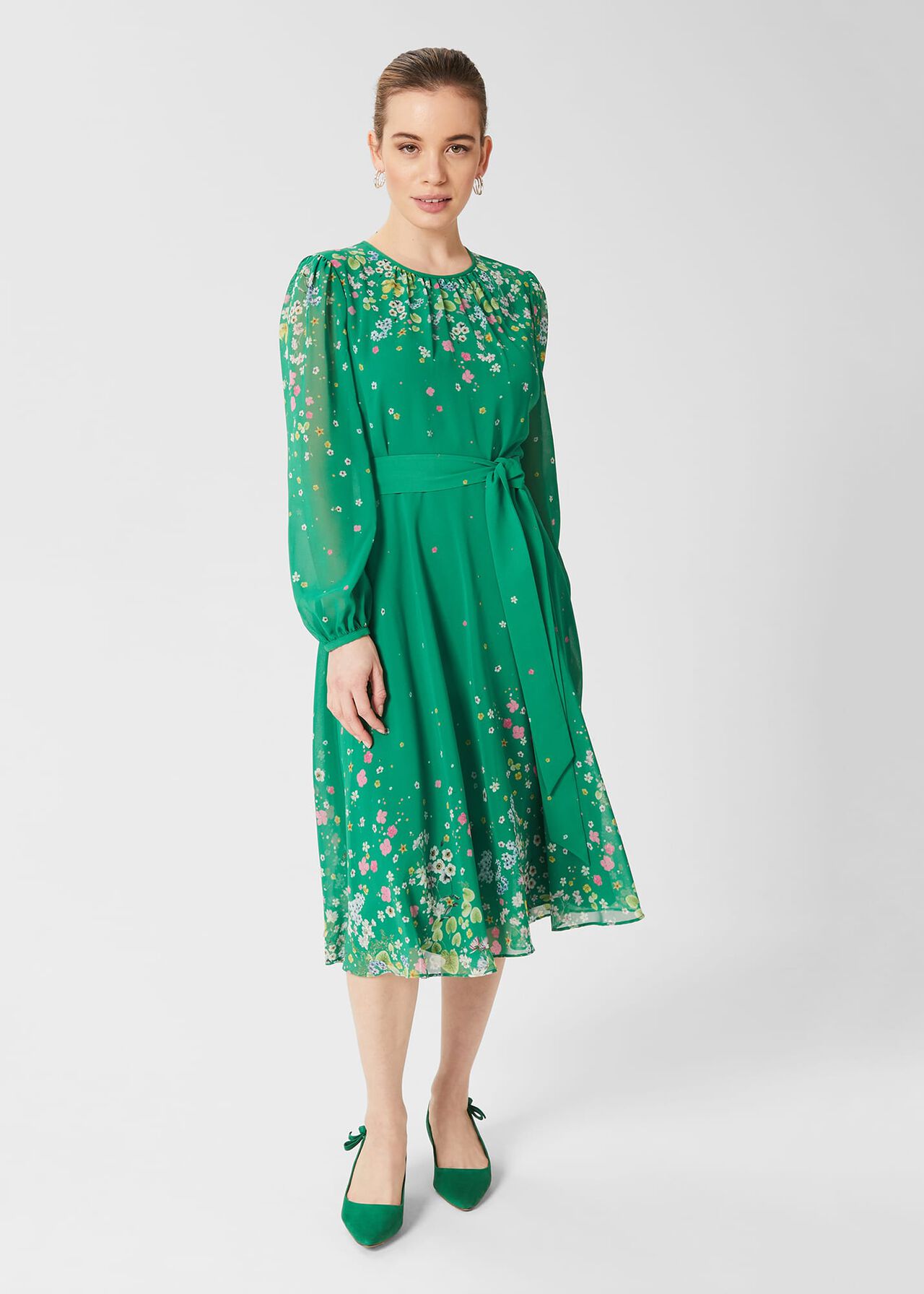 A woman wearing a green floral dress with long sheer sleeves, a crew neckline, and a tie waist, standing against a plain background.