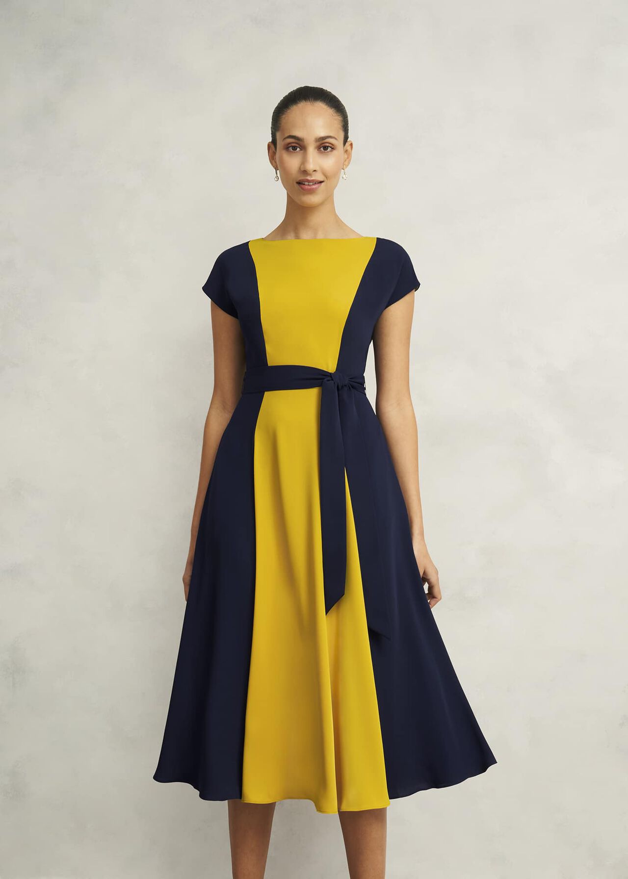 A woman wearing a navy blue and yellow color-block midi dress with a boat neckline, short cap sleeves, and a fabric belt tied at the waist.