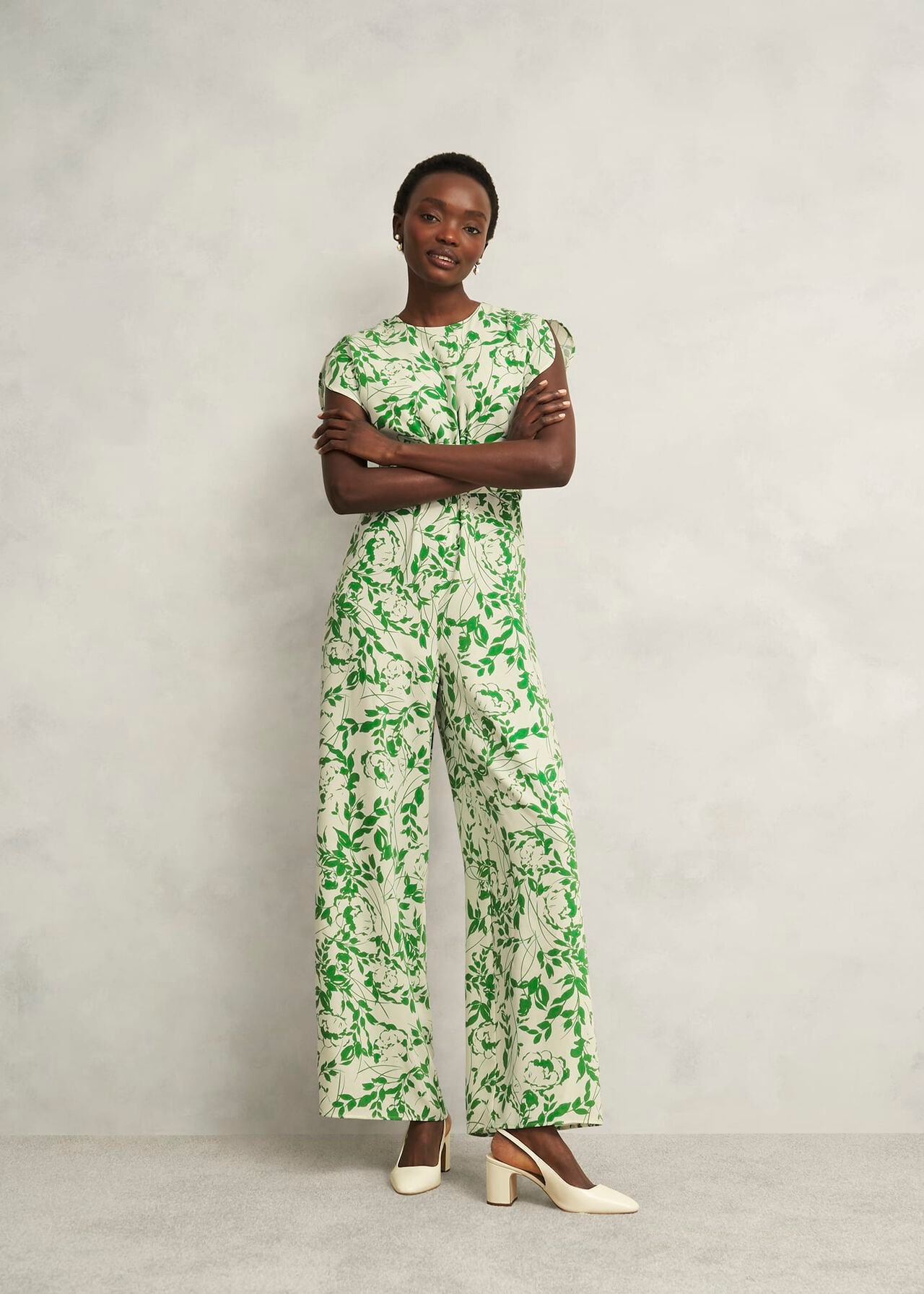 A woman wearing a cream jumpsuit with green floral print, crew neckline, short puff sleeves, and wide-leg pants.