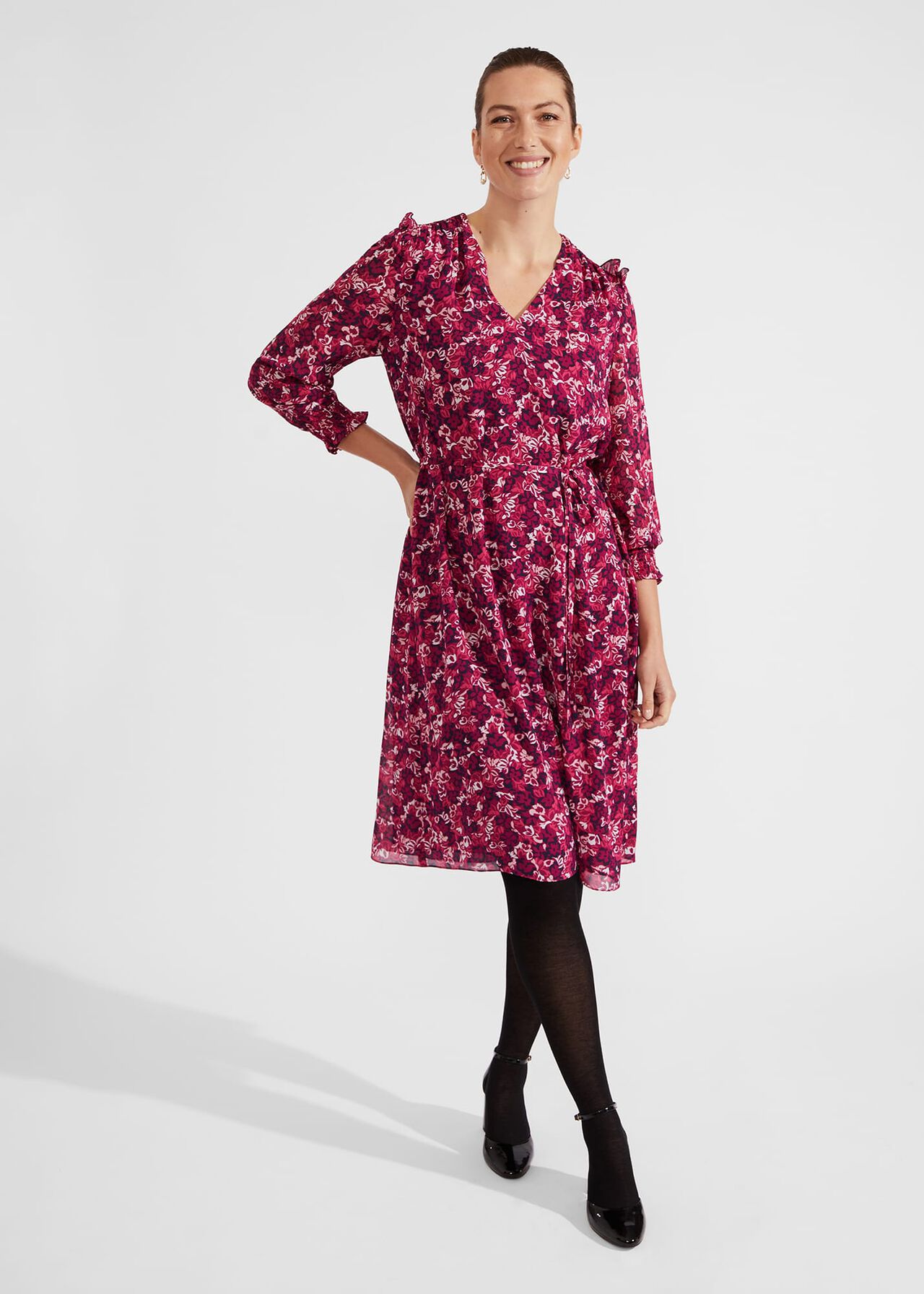 A woman wearing a pink floral midi dress with long sleeves, V-neckline, and ruffle shoulder details.