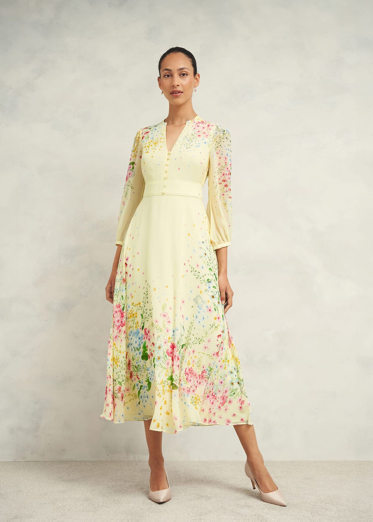 A woman wearing a cream-colored, floral print petite Emma Dress with long puffed sleeves, V-neckline, and midi length, paired with nude heels.