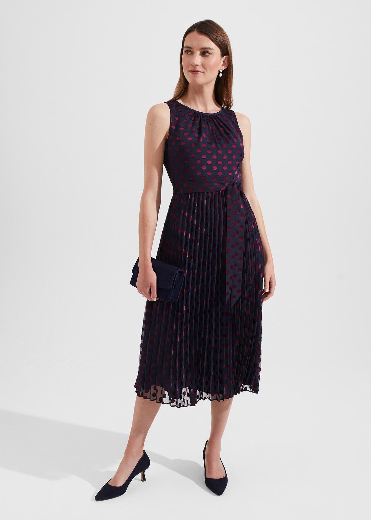 A woman wearing a navy blue sleeveless midi dress with red polka dots, pleated skirt, and a crew neckline, holding a navy clutch, with black heels.