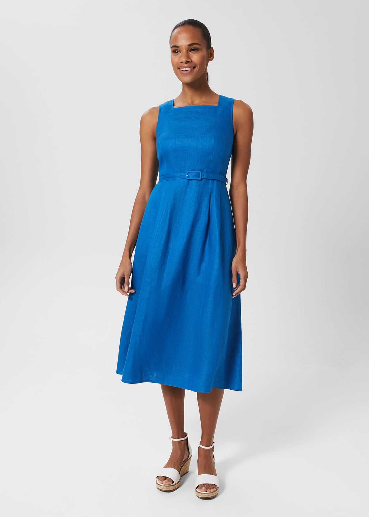 A woman wearing a bright blue sleeveless linen dress with a square neckline, a matching belt at the waist, and a midi length.
