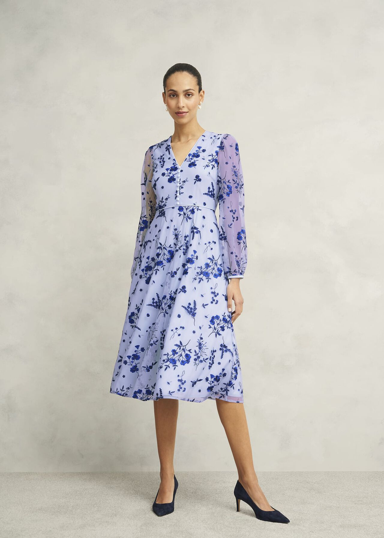 A woman wearing a light purple midi dress with blue floral embroidery, long sheer sleeves, and a V-neckline, standing against a plain background.
