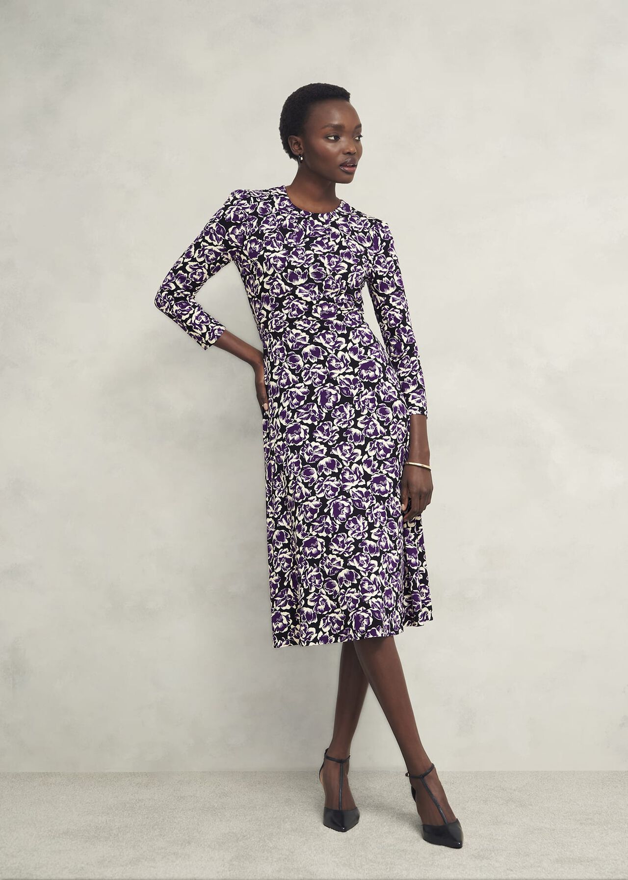 A woman wearing a purple and white floral jersey dress with long sleeves and a crew neckline, standing against a plain background.