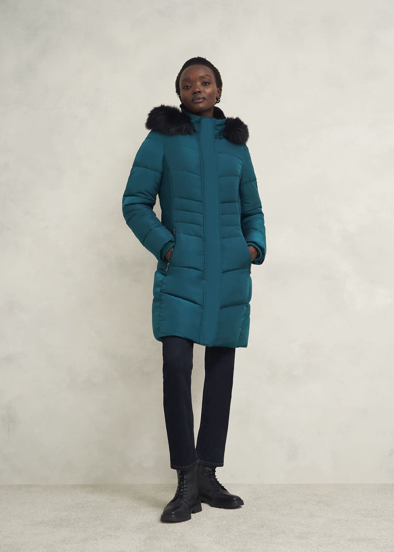 A woman wearing a teal hooded puffer coat with black faux fur trim on the hood, standing against a plain background.