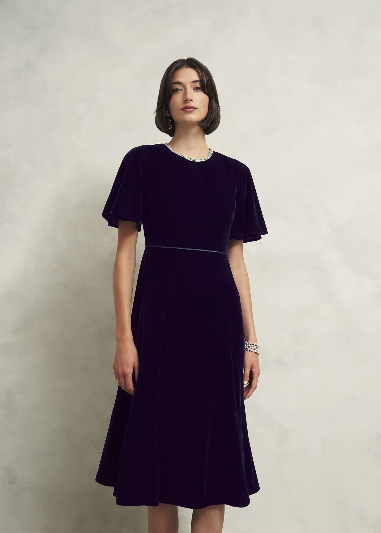 A woman wearing a navy blue velvet dress with short flutter sleeves, a crew neckline, and a fitted waist, standing against a plain background.