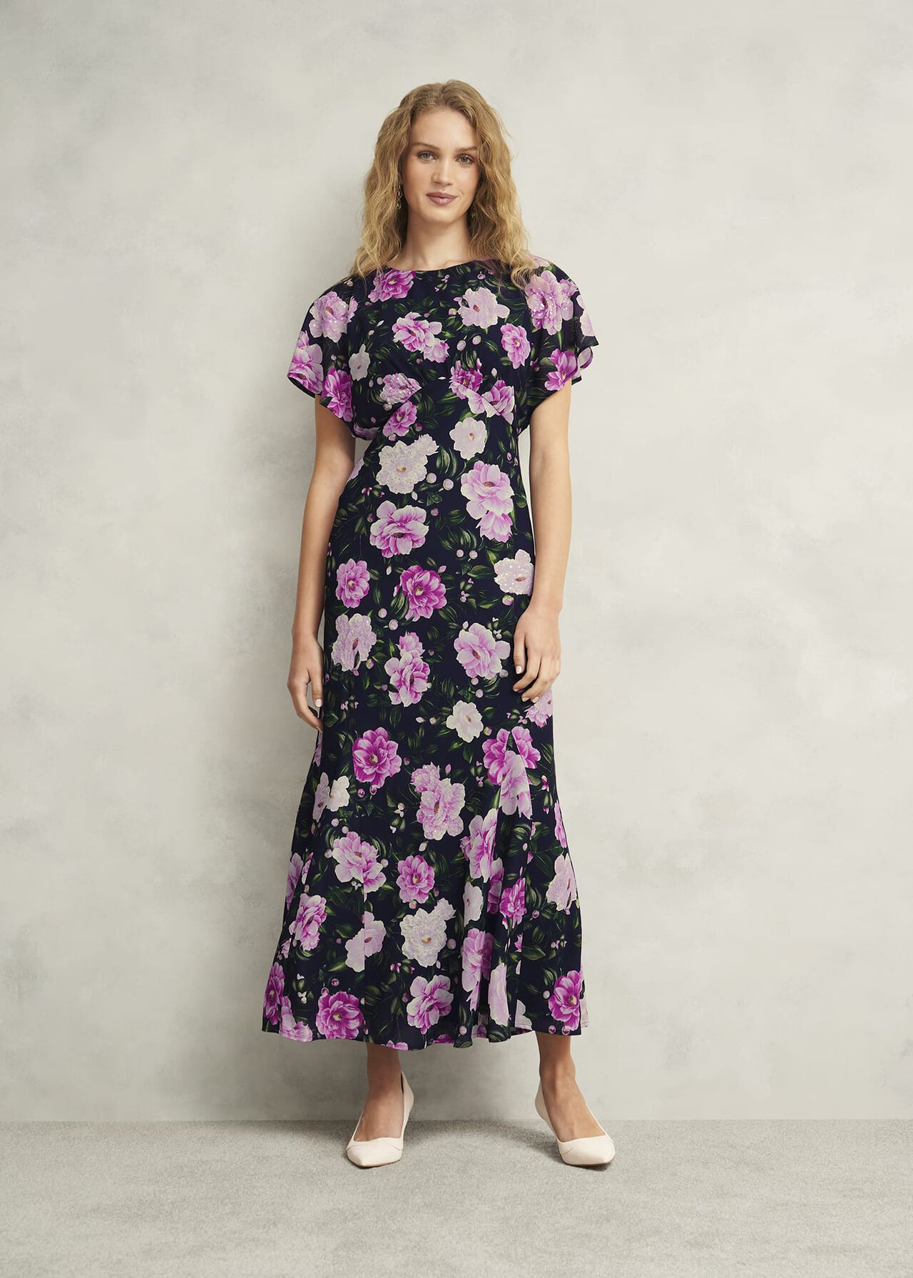 A woman wearing a black floral dress with pink, purple, and white flowers, crew neckline, short flutter sleeves, and midi length.