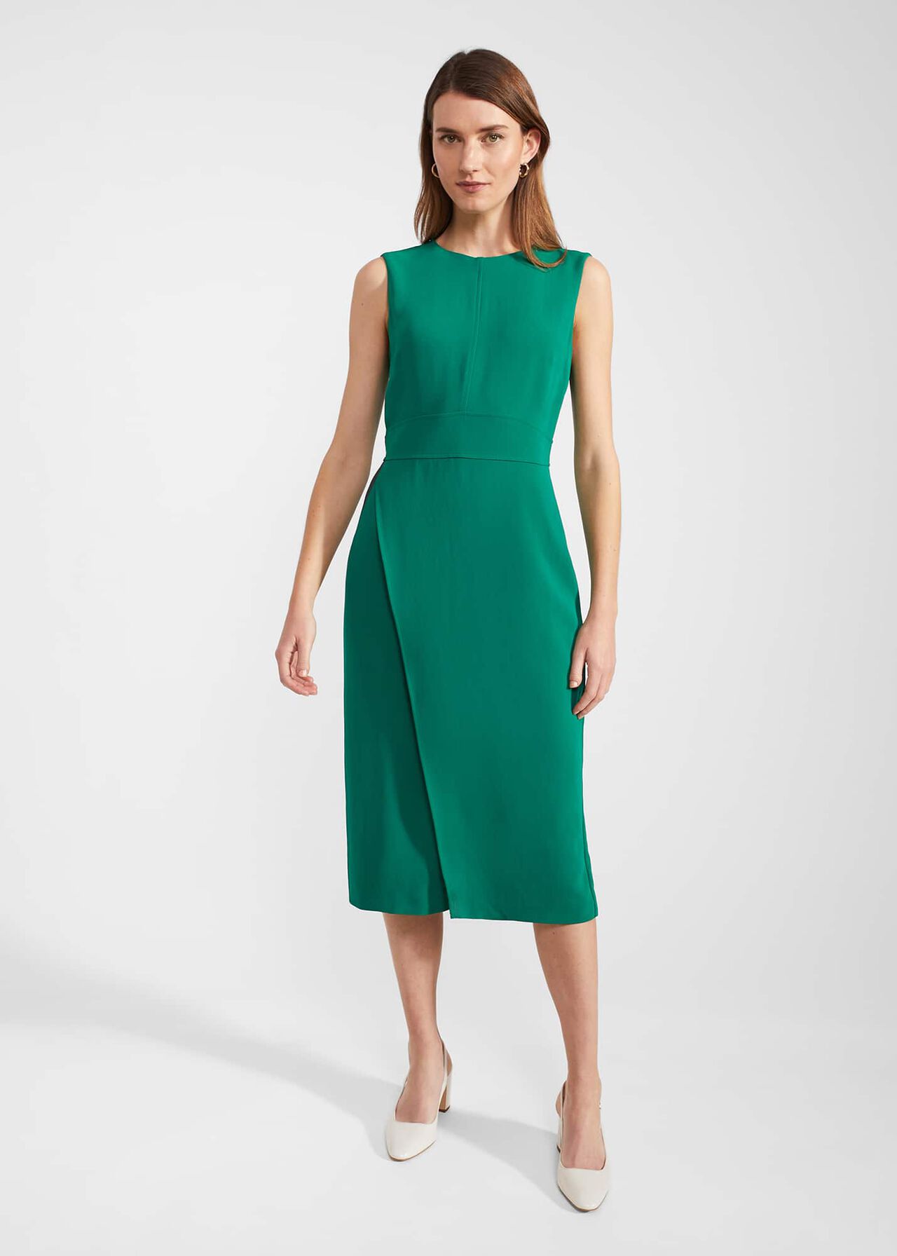 A woman wearing a green sleeveless midi dress with a crew neckline, fitted waist, and front slit, paired with white heels.