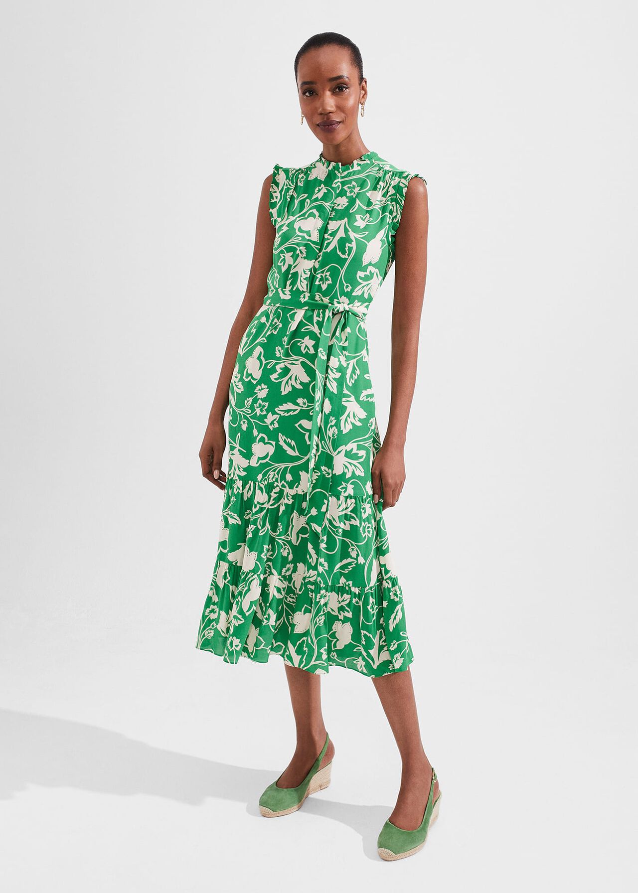 A woman wearing a green sleeveless midi dress with a white floral pattern, mandarin collar, and waist tie, standing against a plain background.