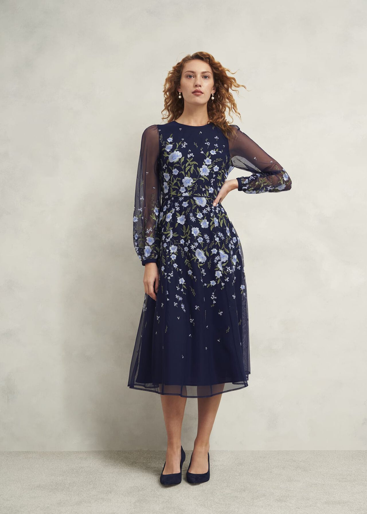 A woman wearing a navy blue embroidered dress with floral patterns, sheer long sleeves, and a crew neckline.
