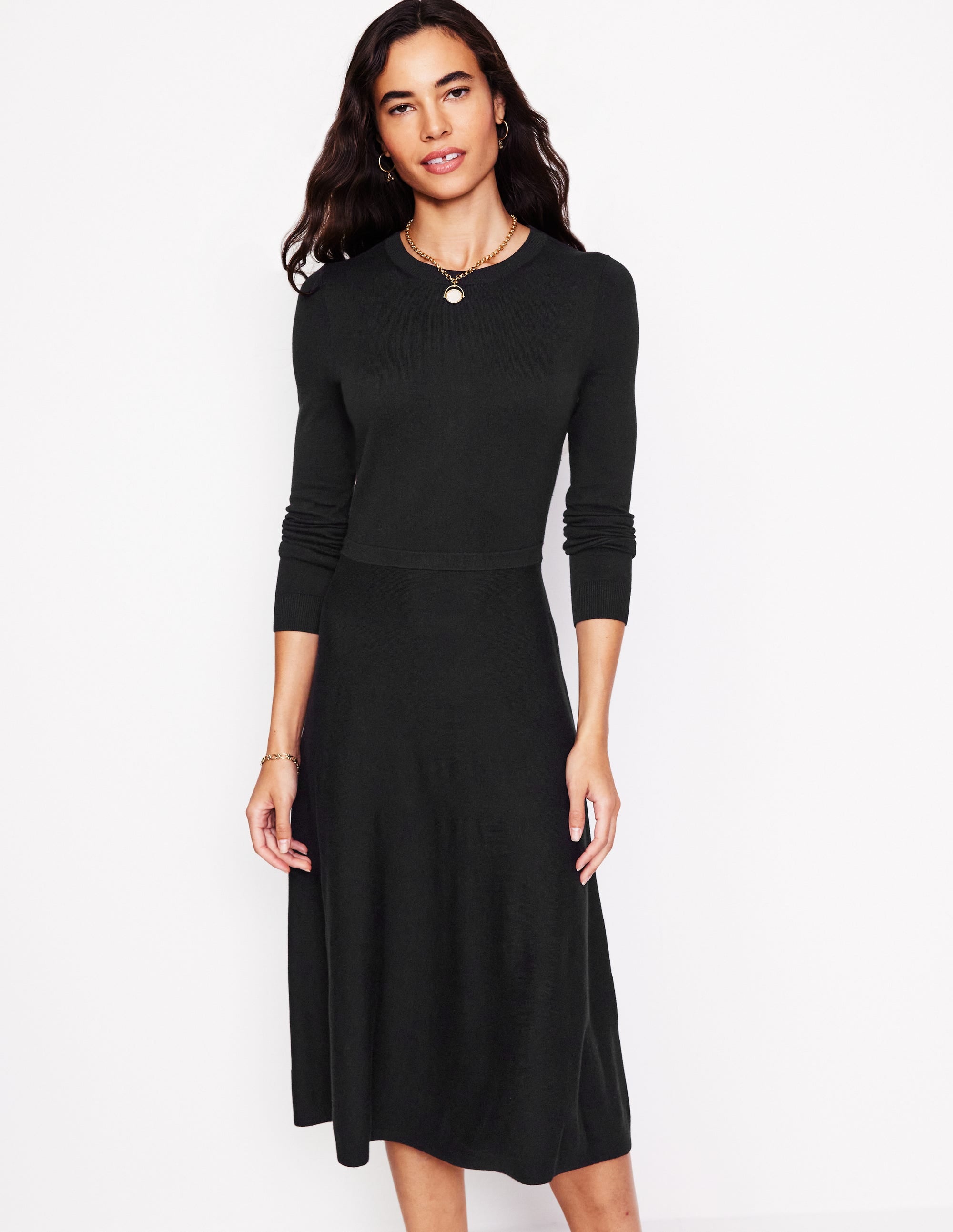 A woman wearing a black knitted midi dress with long sleeves, a round neckline, and a flared skirt, standing against a plain background.