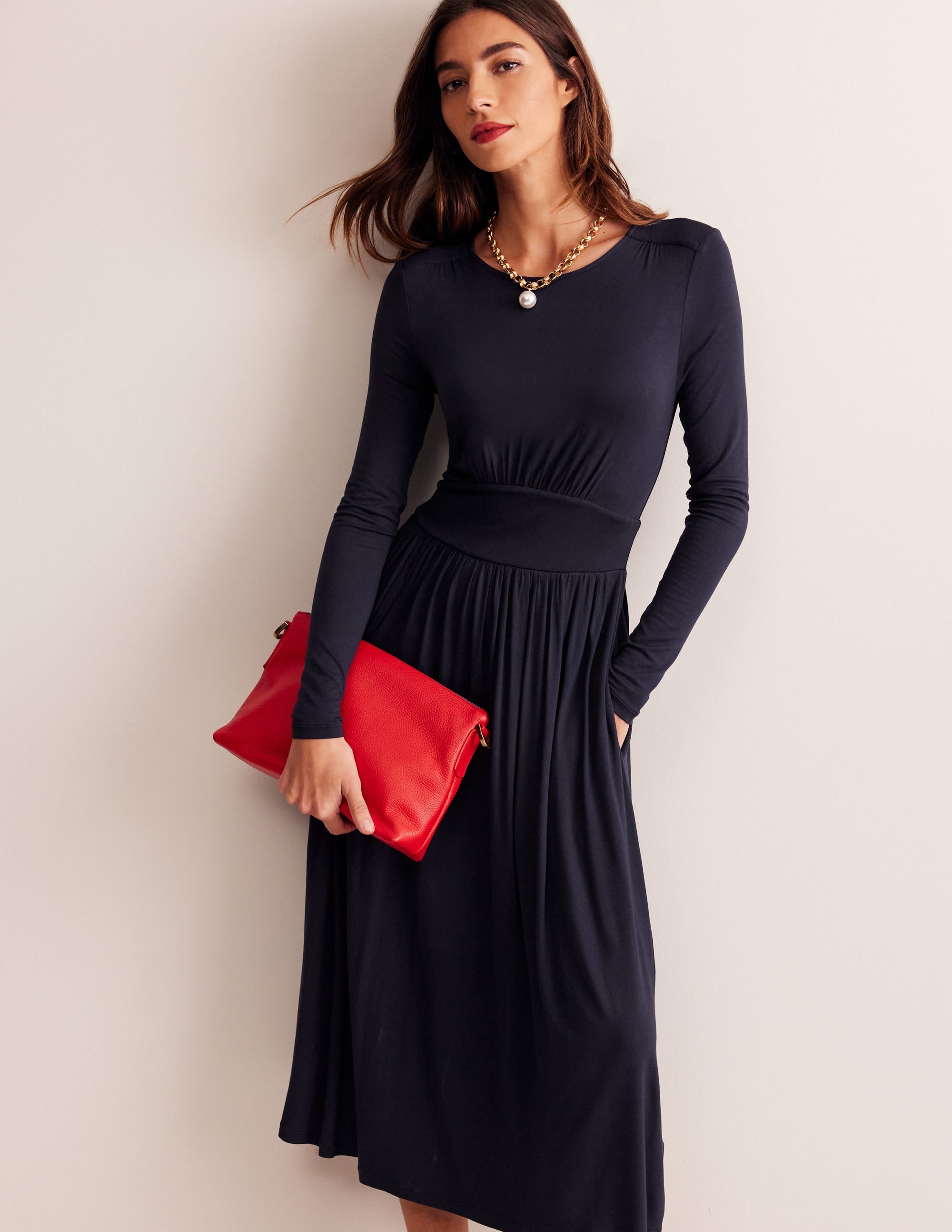 A woman wearing a navy long sleeve jersey dress with a gathered skirt and waist panel, paired with a gold chain necklace and holding a red clutch.