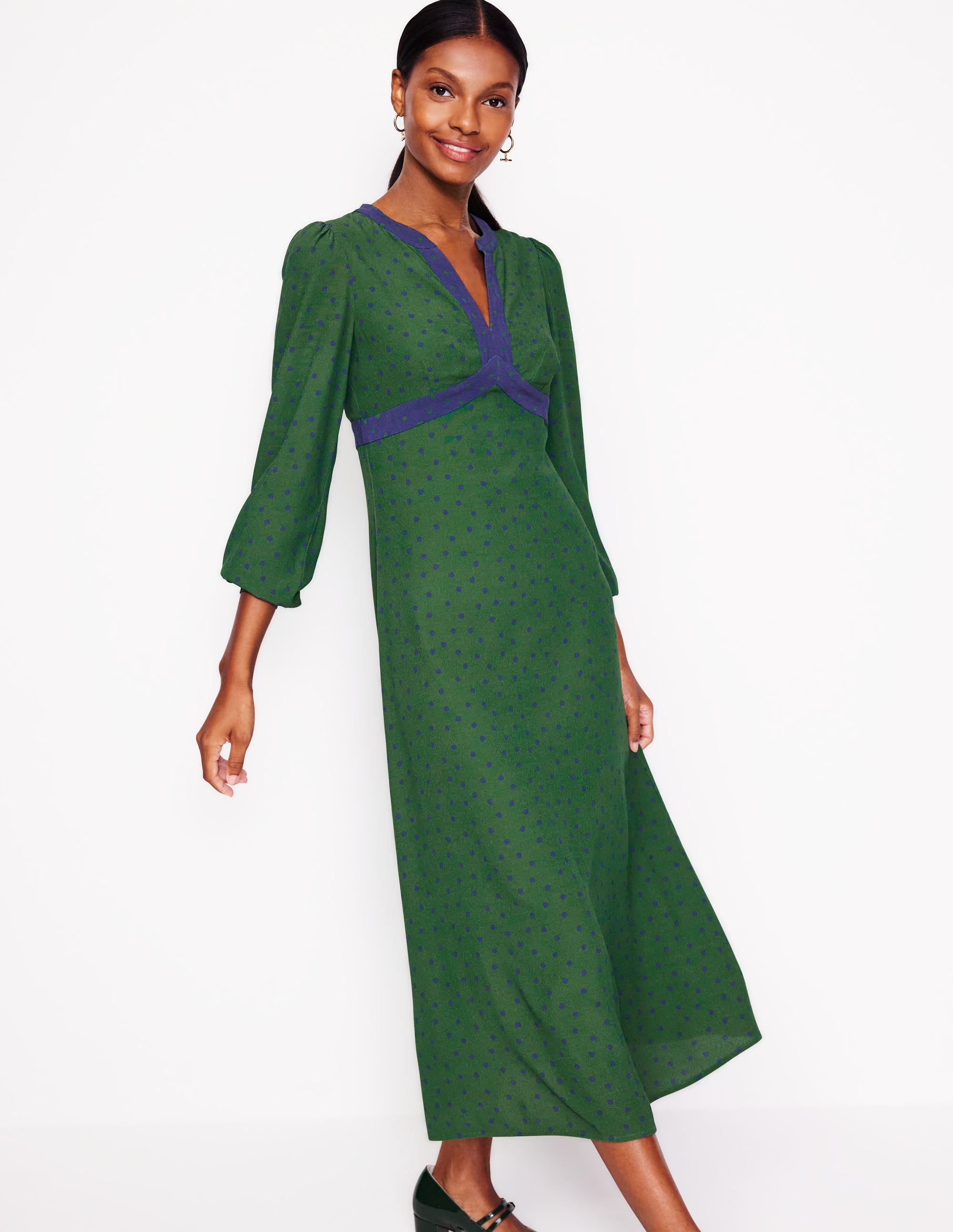 A woman wearing a green midi dress with blue spot border print, long sleeves, and a fitted silhouette, standing against a white background.