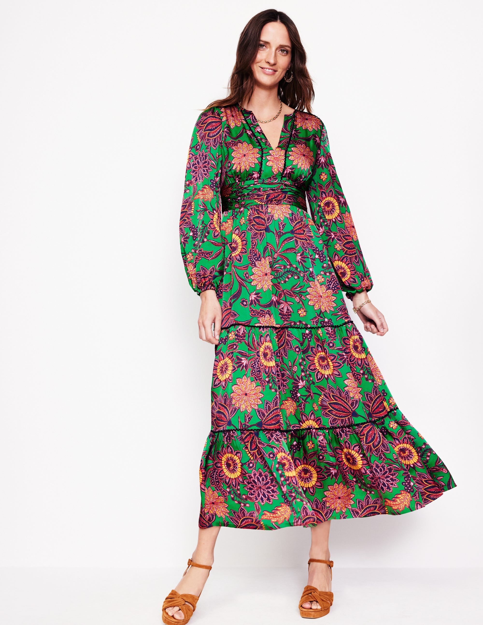 A woman wearing a green satin maxi dress with floral and ornate bud patterns, long sleeves, and tiered layers.