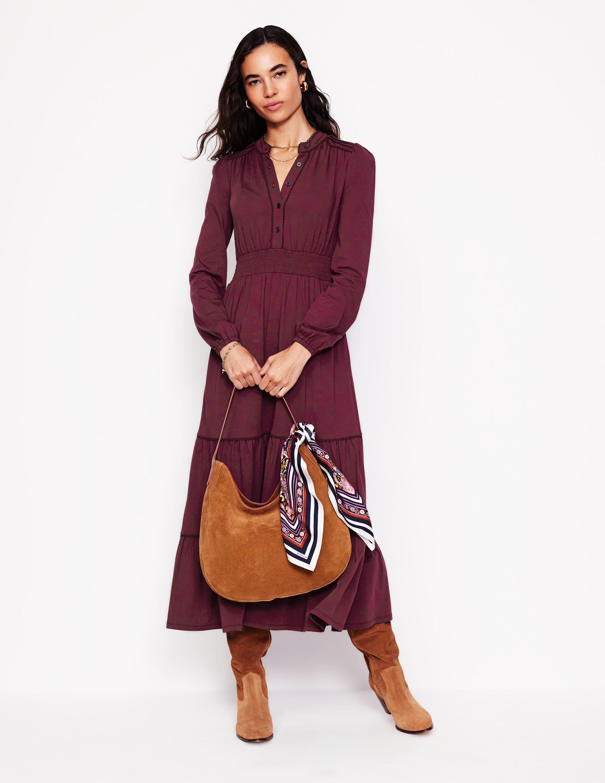 A woman wearing a Deep Wisteria colored midi dress with long sleeves, tiered skirt, and shirred waist, holding a brown handbag with a colorful scarf.