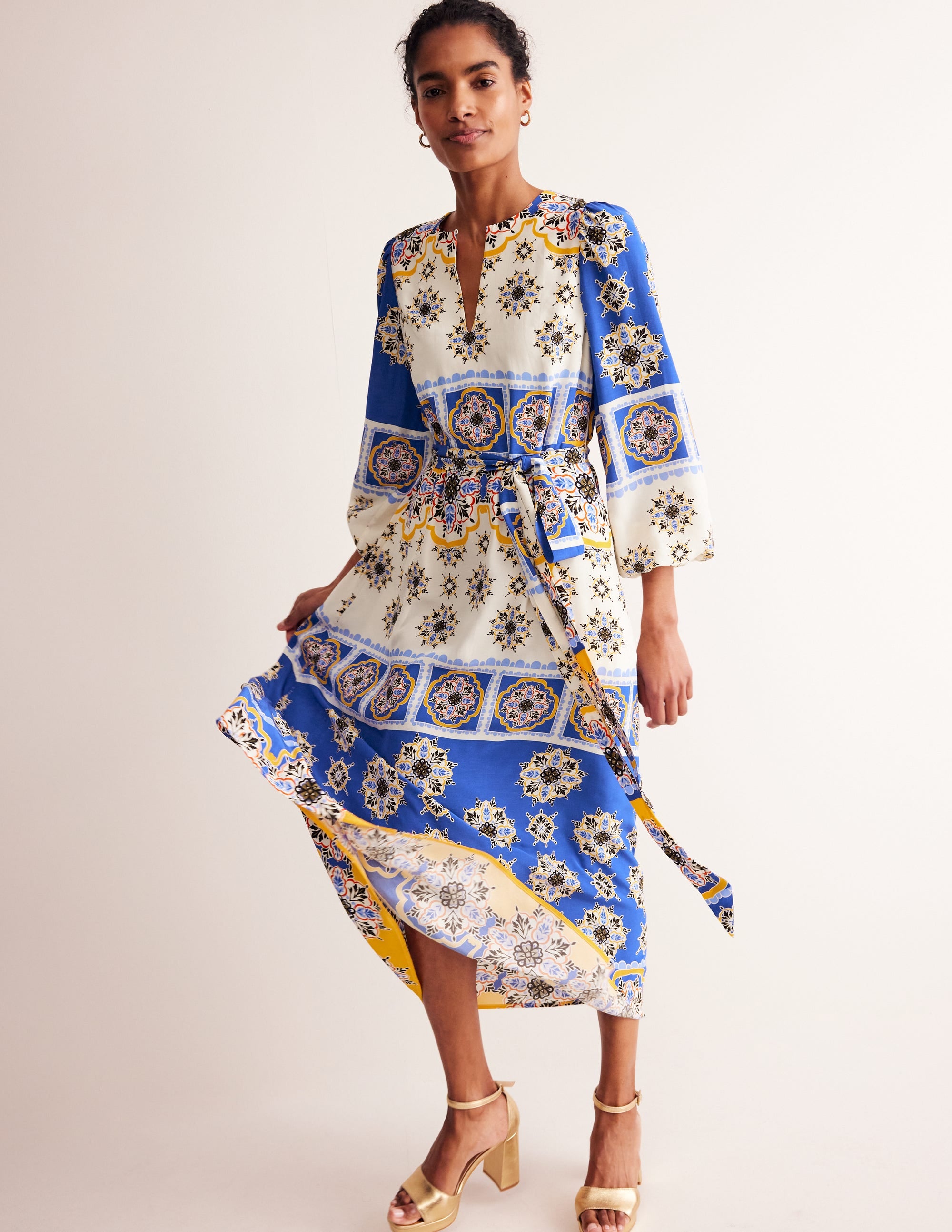 A woman wearing a long, colorful mosaic-patterned maxi dress with blouson sleeves, a fabric belt, and a keyhole neckline.