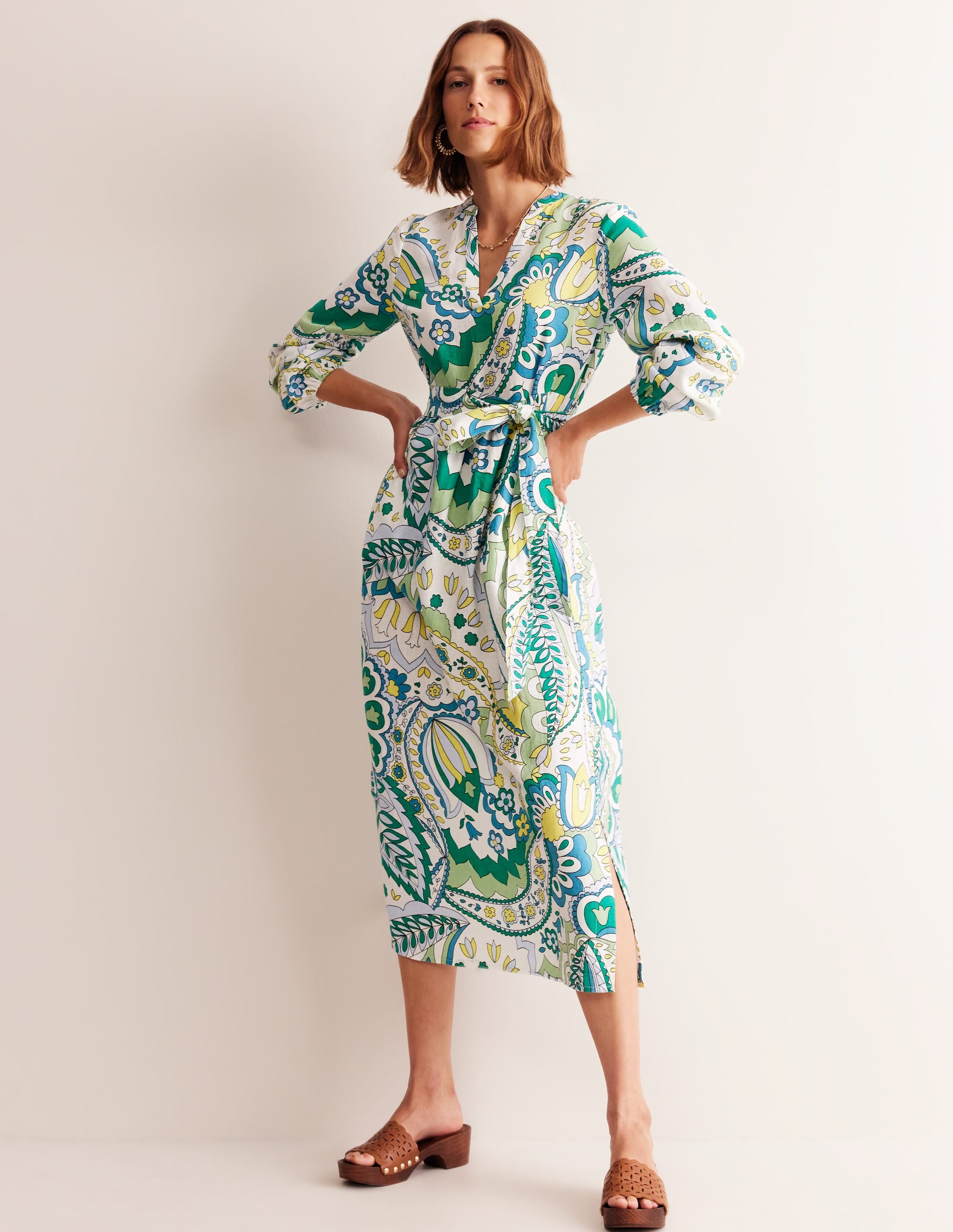 A woman wearing a long linen maxi dress with a foliage paisley pattern in shades of emerald, green, yellow, and white, featuring a notch neckline, blouson sleeves, and side splits.