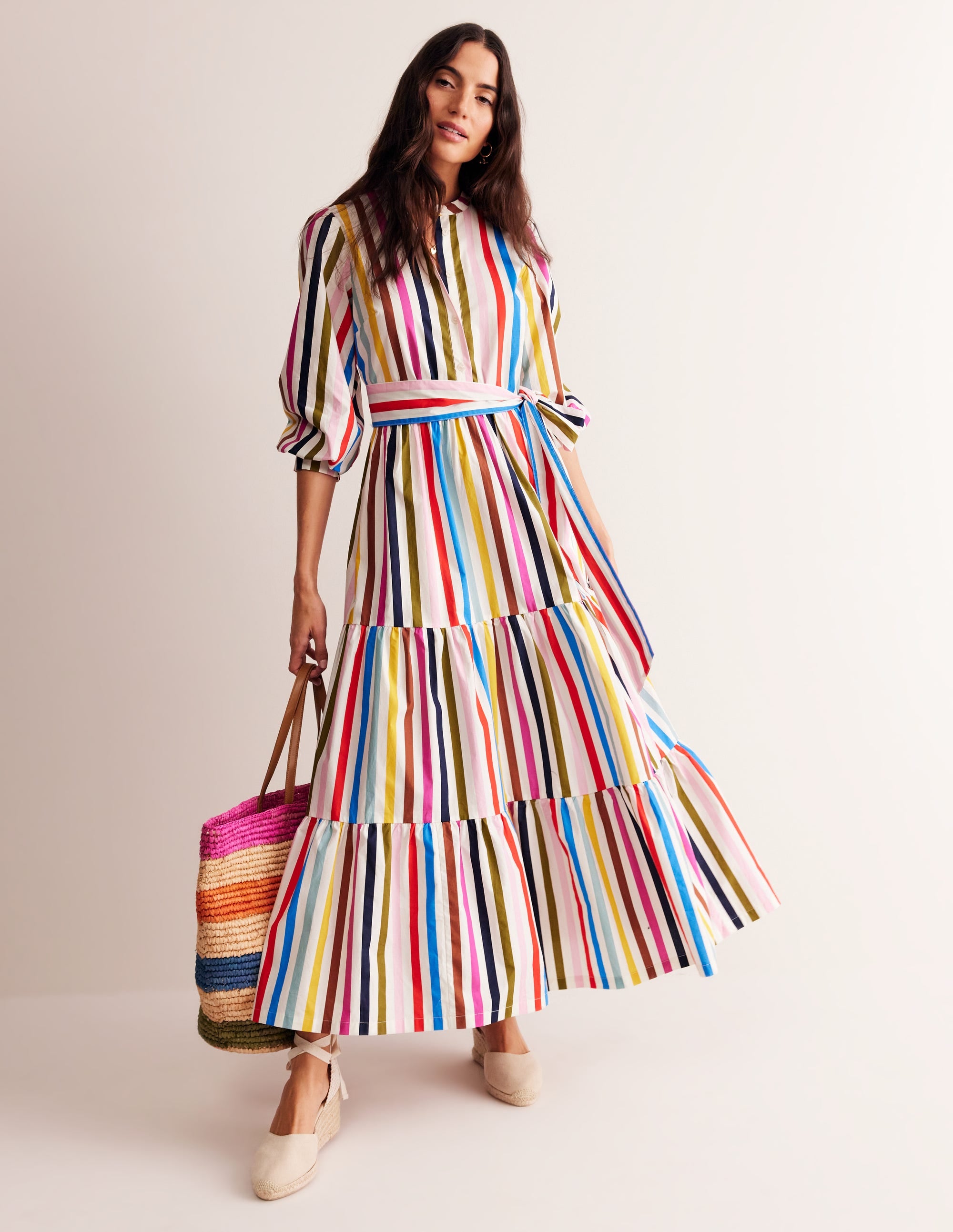 A woman wearing a multicolour striped maxi dress with long sleeves, a rounded neckline, and a self-tie waist, standing against a plain background.
