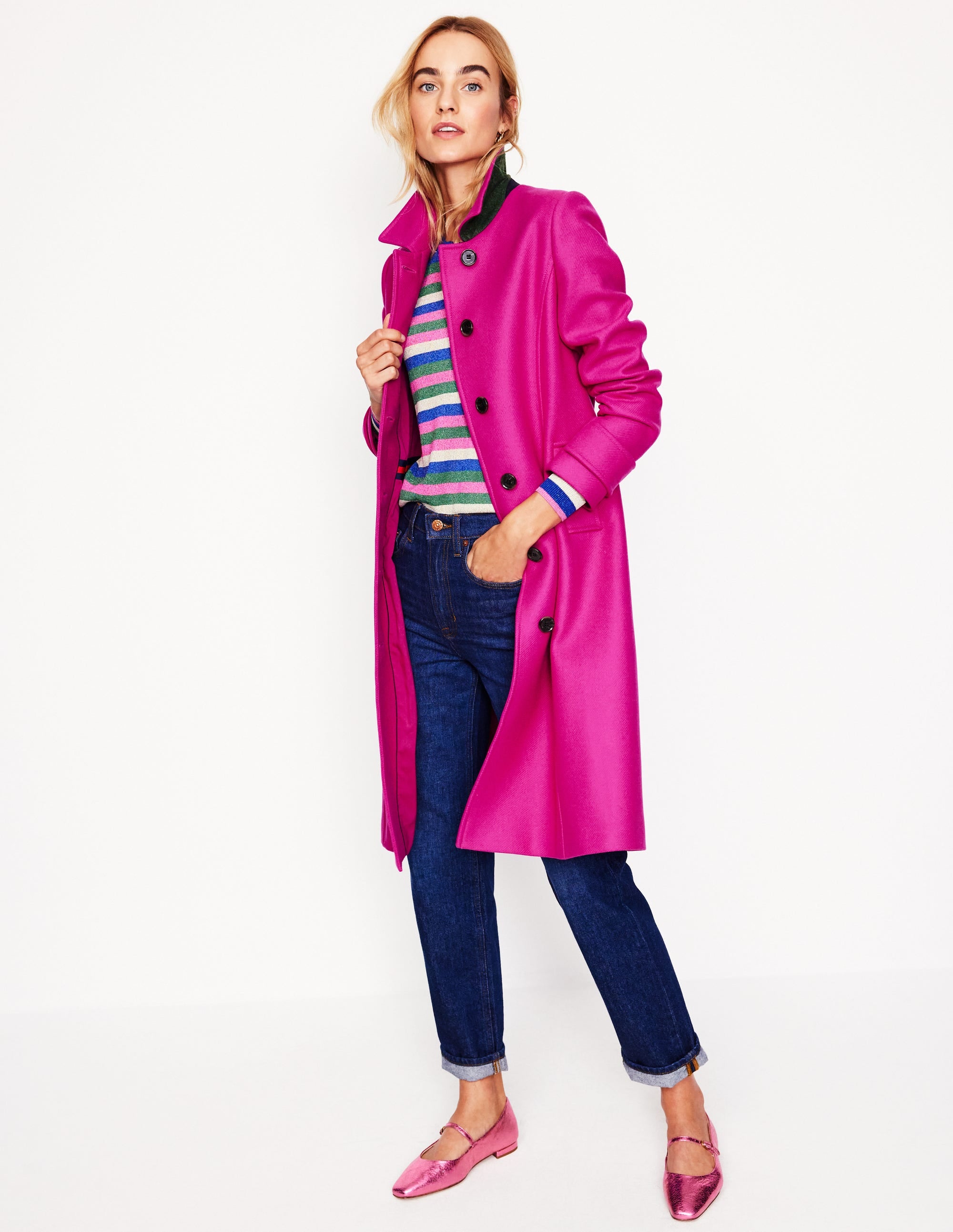A woman wearing a bright pink wool blend coat with a tailored fit, princess seams, a smart collar, and button closure, paired with a colorful striped sweater, jeans, and pink shoes.