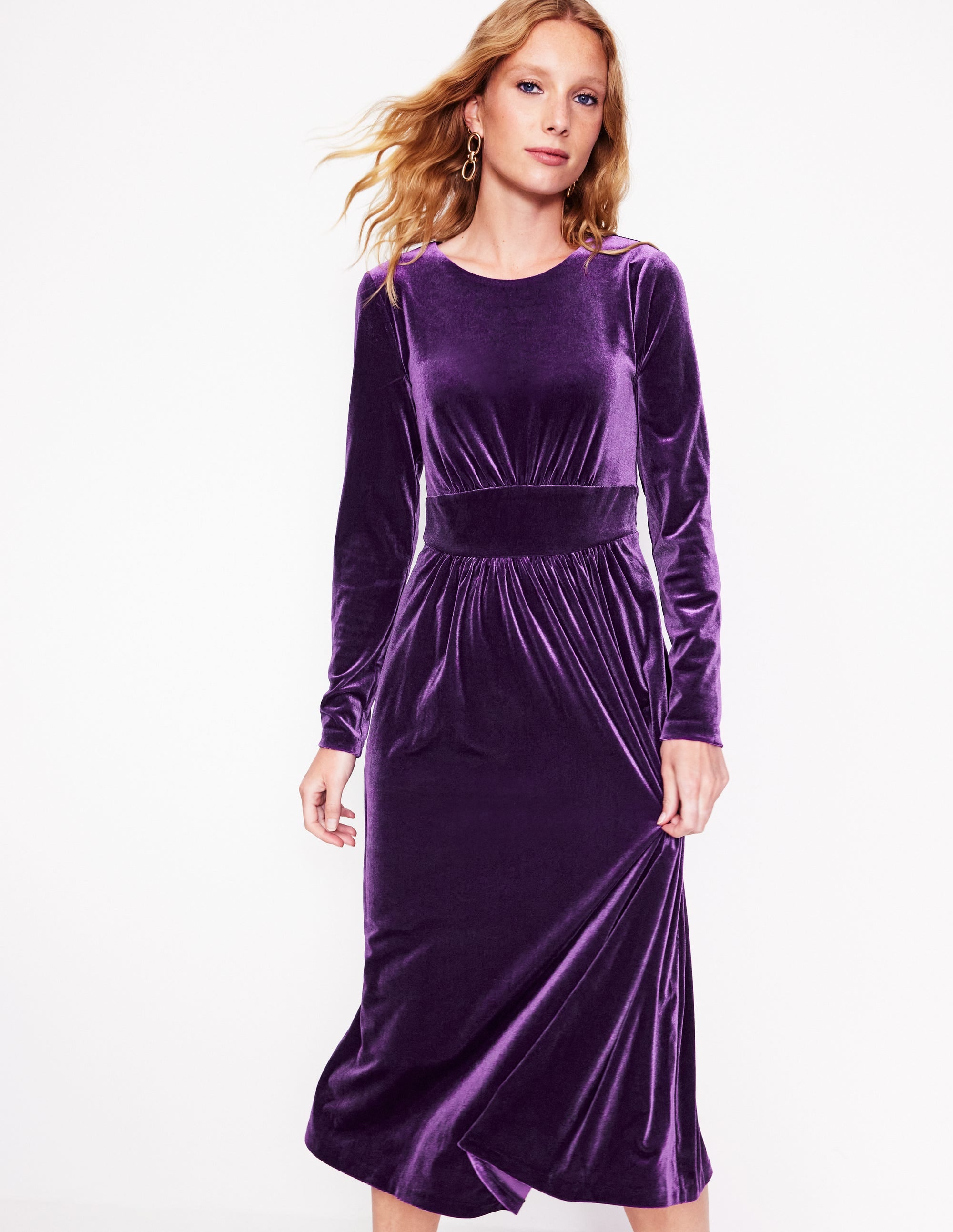 A woman wearing a purple velvet midi dress with long sleeves, a ruched bodice, and a fitted waist.